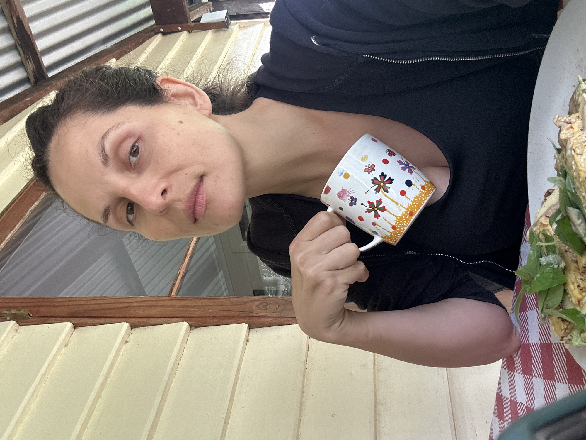 A woman is sitting on a porch with a red and white checkered tablecloth in front of her. She is holding a mug with a floral design. There is a plate of food on the table. The porch has a wooden railing and a corrugated iron roof. The woman is wearing a black zip-up jacket over a black top. The house is a weatherboard house painted cream.