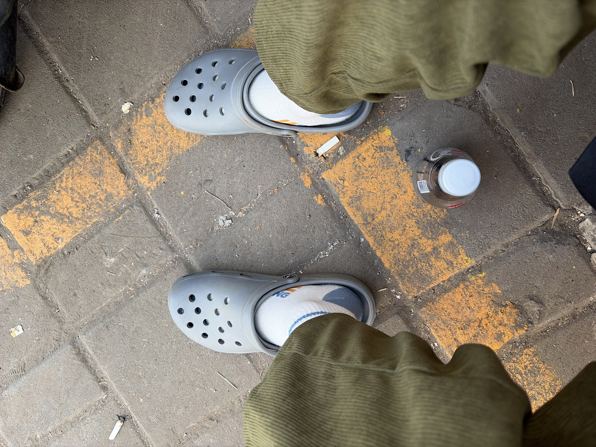 A person wearing grey Crocs and olive green corduroy pants is sitting on a paved surface marked with yellow lines. A bottle with a white cap is on the ground next to the person's right foot. Two cigarette butts are visible on the ground near the person's left foot. The scene appears to be outdoors, possibly in a public area within Secang, Indonesia, given the presence of paved walkways and common urban debris like cigarette butts.