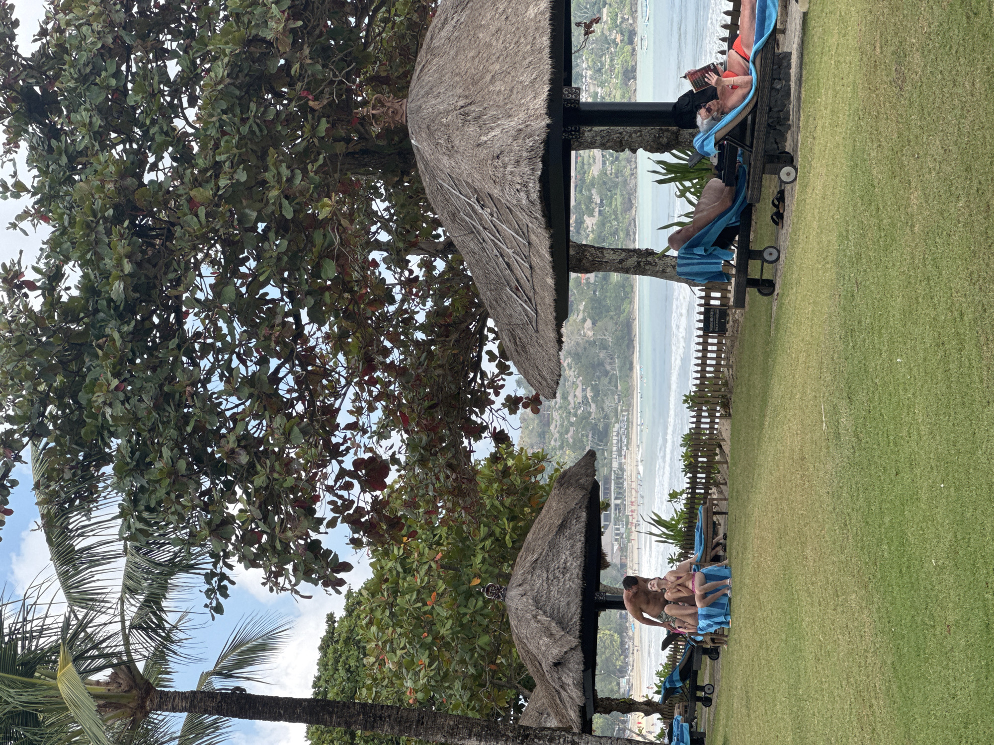 This is a photograph taken outdoors in Kuta Selatan, Indonesia, near a beach. The image shows a grassy area with several thatched-roof umbrellas providing shade. In the foreground, on the right side, two individuals are relaxing on lounge chairs. One person, wearing red swimwear, is reading a book. The other person, mostly obscured by the umbrella, is also on a lounge chair. On the left side, under a separate umbrella, a man is standing and leaning towards two women who are sitting on lounge chairs. They are all wearing swimwear and appear to be interacting. Beyond the lounge chairs, a wooden fence separates the grassy area from a beach with people and umbrellas. The ocean is visible with waves and several boats in the distance. Lush green trees and palm trees are abundant in the background, with a hazy, hilly landscape behind them. The sky is partly cloudy.