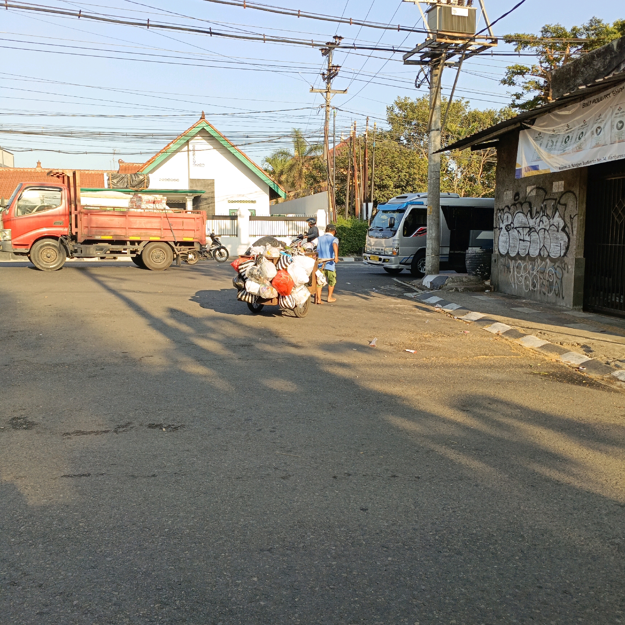 In a street scene in Salatiga, Indonesia, a red flatbed truck is parked on the left side of the asphalt road. The truck is loaded with several stacks of white material, possibly lumber or construction supplies, covered with a black tarp. In the center of the image, a man in a blue t-shirt and green shorts pushes a cart loaded with many bags of various sizes and colors. Behind him, another person on a motorcycle is partially visible. To the right of the cart, a silver Isuzu van is parked. A building with graffiti on its wall and a sign that reads "BIMA POULTRY SHOP" is located on the right side of the road. Overhead, numerous power lines crisscross the clear blue sky. The sun is casting long shadows across the road, indicating it might be late afternoon. The environment appears to be a residential or commercial area with houses and trees visible in the background.