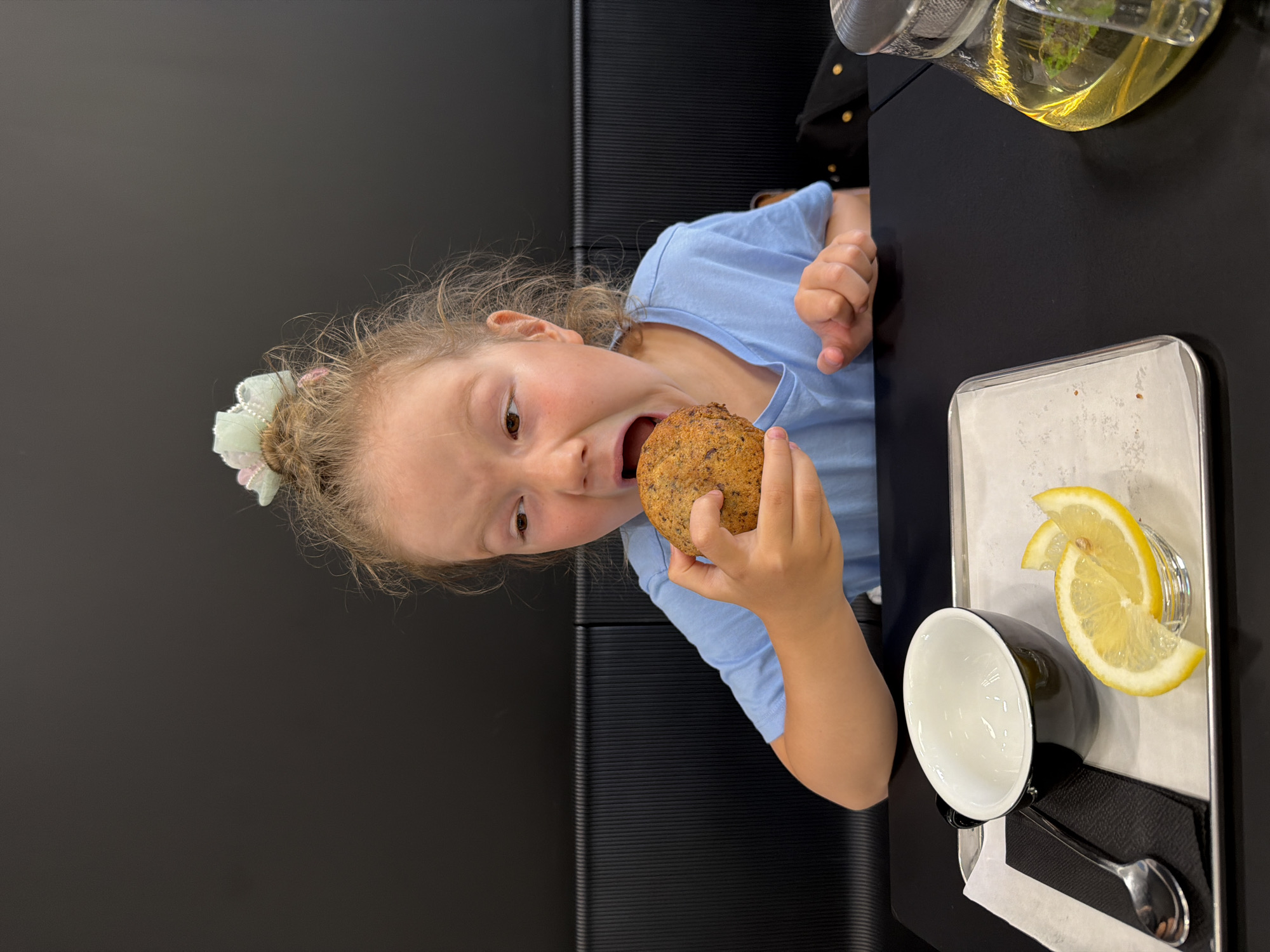 A young girl with blonde hair in a messy bun, adorned with a pastel scrunchie, is pictured in a close-up, medium shot, indoors, at a table. She is wearing a light blue t-shirt and is in the process of biting into a round, textured baked good, likely a muffin or scone, held in her right hand. Her mouth is open, and her eyes are directed towards the food. In the foreground, on a silver tray, there is a black teacup with a white interior, a folded black napkin, a silver spoon, and two lemon slices. To the right, a glass teapot containing a yellow liquid and possibly mint leaves is visible. The background consists of a dark, textured wall and black seating. The image appears to have been taken at a cafe or restaurant, possibly in Izhevsk, Russia, given the context of the prompt, though there are no explicit visual cues within the image to confirm this location. The lighting is even, suggesting an indoor setting with artificial light.