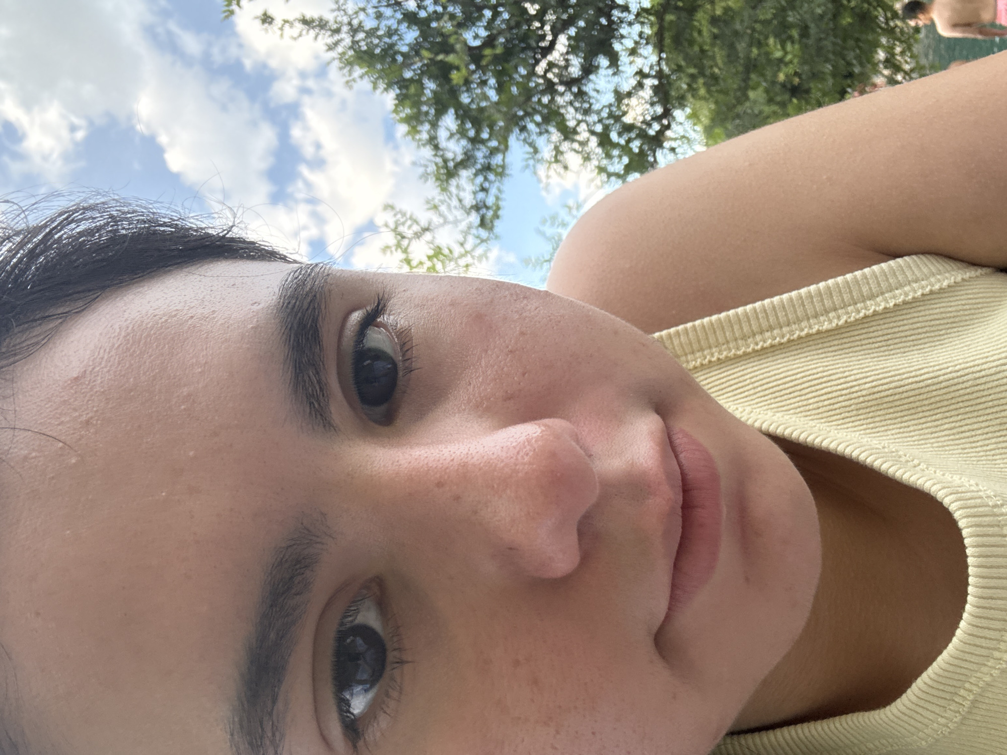 The image is a close-up selfie of a person with dark hair and eyes, wearing a yellow ribbed top. The background shows a blue sky with white clouds and green trees. The setting appears to be outdoors, possibly in a park or natural area. There's a partial view of someone else in the background, possibly wearing swimwear. The picture was likely taken in Austin, United States of America, as requested.