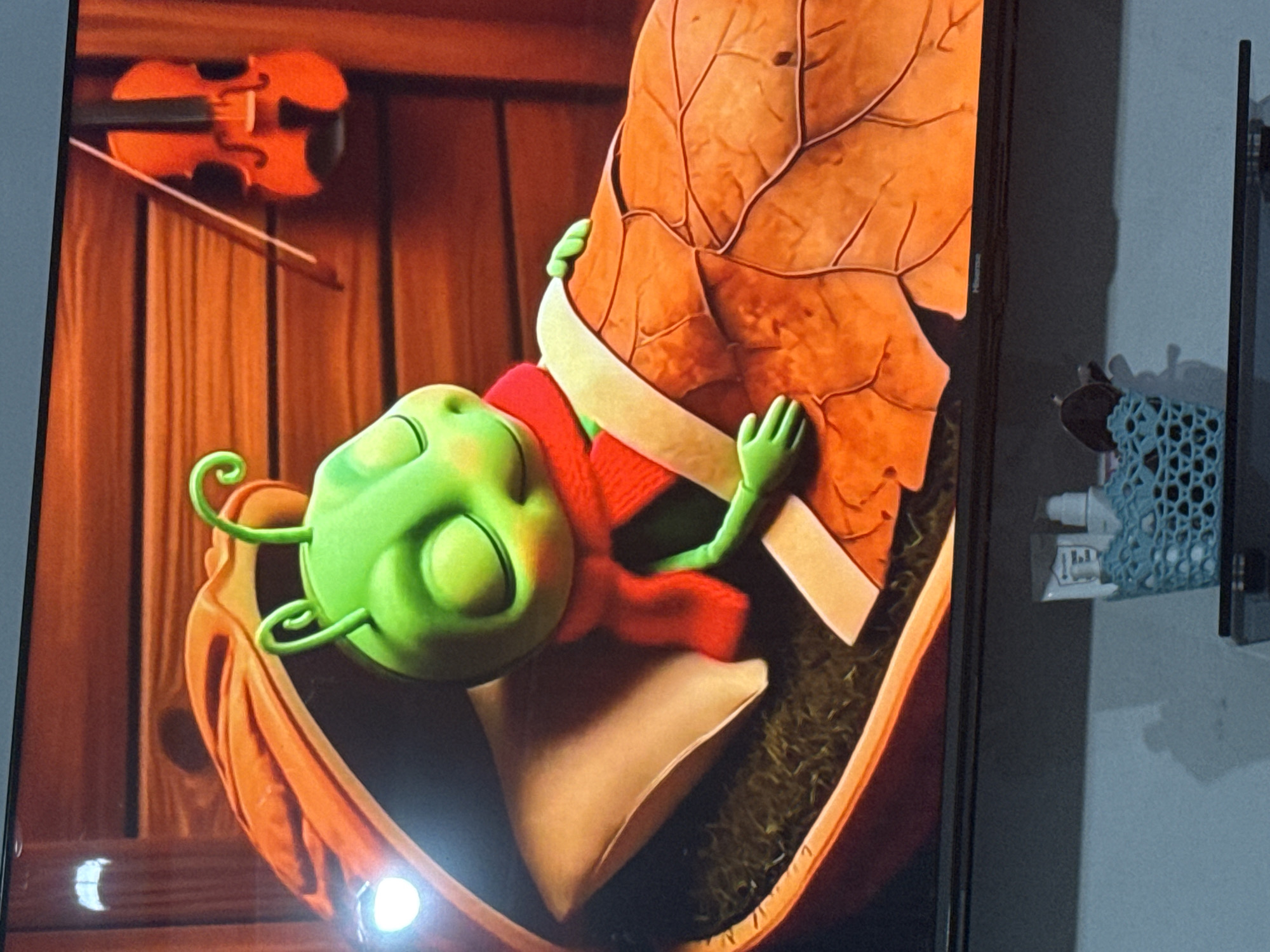 The image shows a television screen displaying a scene from an animated production. The central subject is a green, cartoonish insect character lying in a bed made of what appears to be a large, orange leaf. The insect is wearing a red scarf and is covered by a blanket. It has its eyes closed and a peaceful expression. Behind the insect, a wooden wall is visible, with a miniature orange violin and bow hanging on it. The lighting suggests a warm, indoor environment. The location of Chardara, Uzbekistan, is not depicted or inferable from the image content.