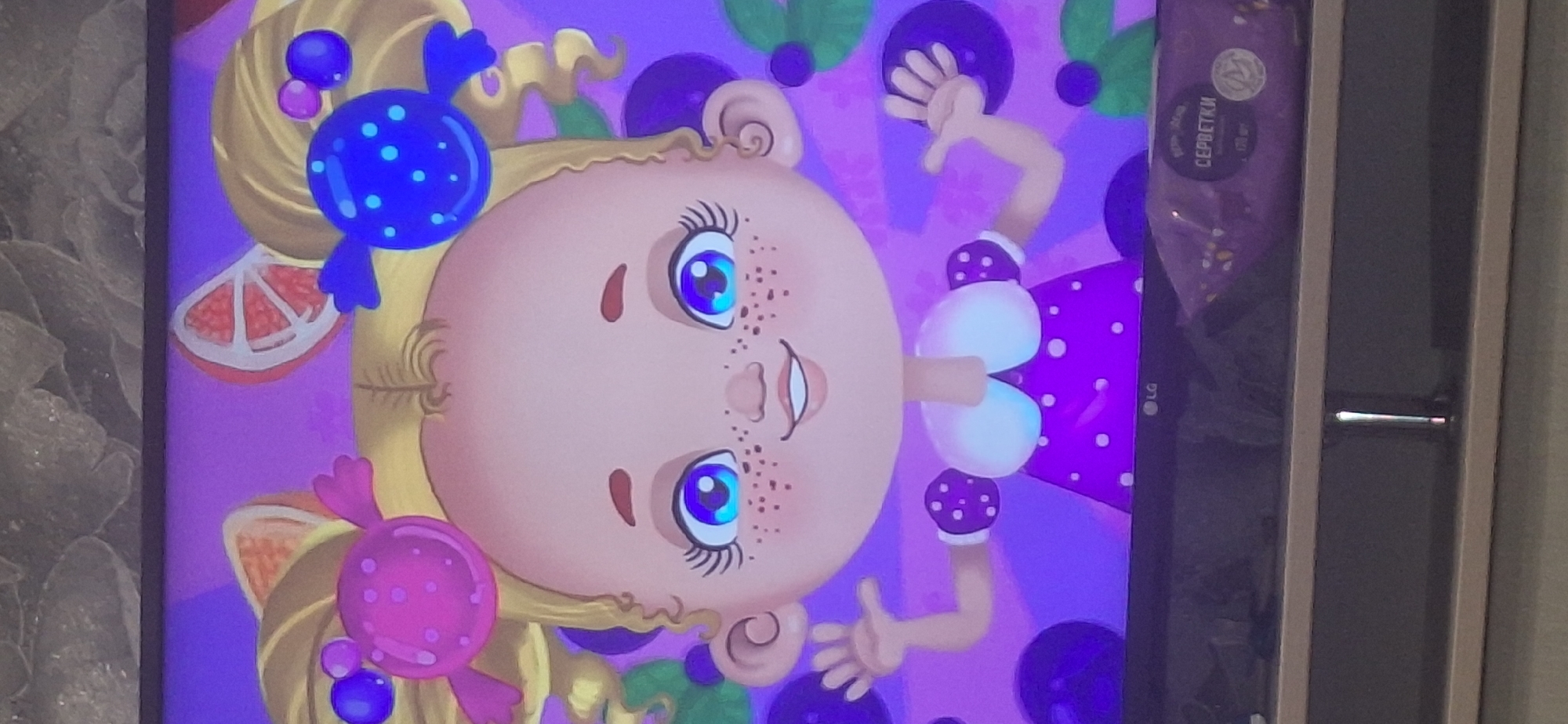 The image shows a close-up of a cartoon drawing of a baby with blonde pigtails adorned with candies, and sliced fruit. The baby has large blue eyes, freckles, and is wearing a purple polka-dot dress. The background is purple with stylized blueberries. Below the cartoon, a television screen is visible, with a portion of a product package featuring the word "СЕРБЕТКИ" (servetki, meaning napkins in Russian) on the bottom right. The television is resting on a stand with shelves. The setting is indoors, likely a home or business, with a grainy texture in the background suggesting a wall covering. There is no discernible location in Bila Tserkva, Ukraine, or any indication of a specific city within the image itself.