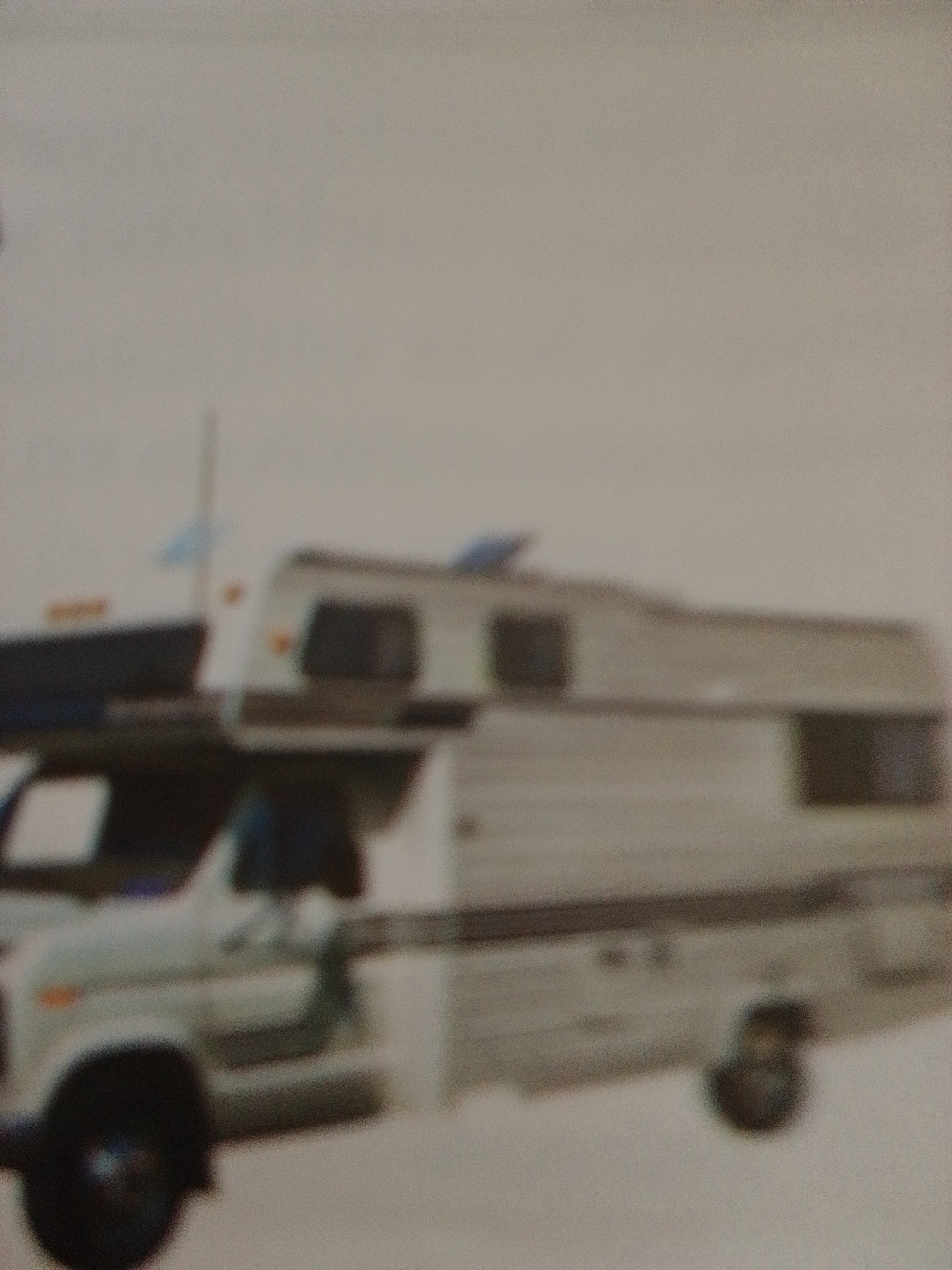 In La Tagua, Venezuela, a blurred image shows a white recreational vehicle (RV) parked outdoors. The RV is the primary subject, taking up a significant portion of the frame. Its body appears to be white with a dark stripe running along its side. The front of the RV, including a portion of the cab, is visible on the left, featuring a black tire. Above the RV, a faint blue object, possibly a flag or antenna, is discernible. The background is a muted, light gray, suggesting an overcast sky or a plain, unadorned environment. No people are clearly visible, and no specific actions or interactions are depicted. The overall image quality is poor, characterized by a significant blur and graininess, making precise details difficult to ascertain.