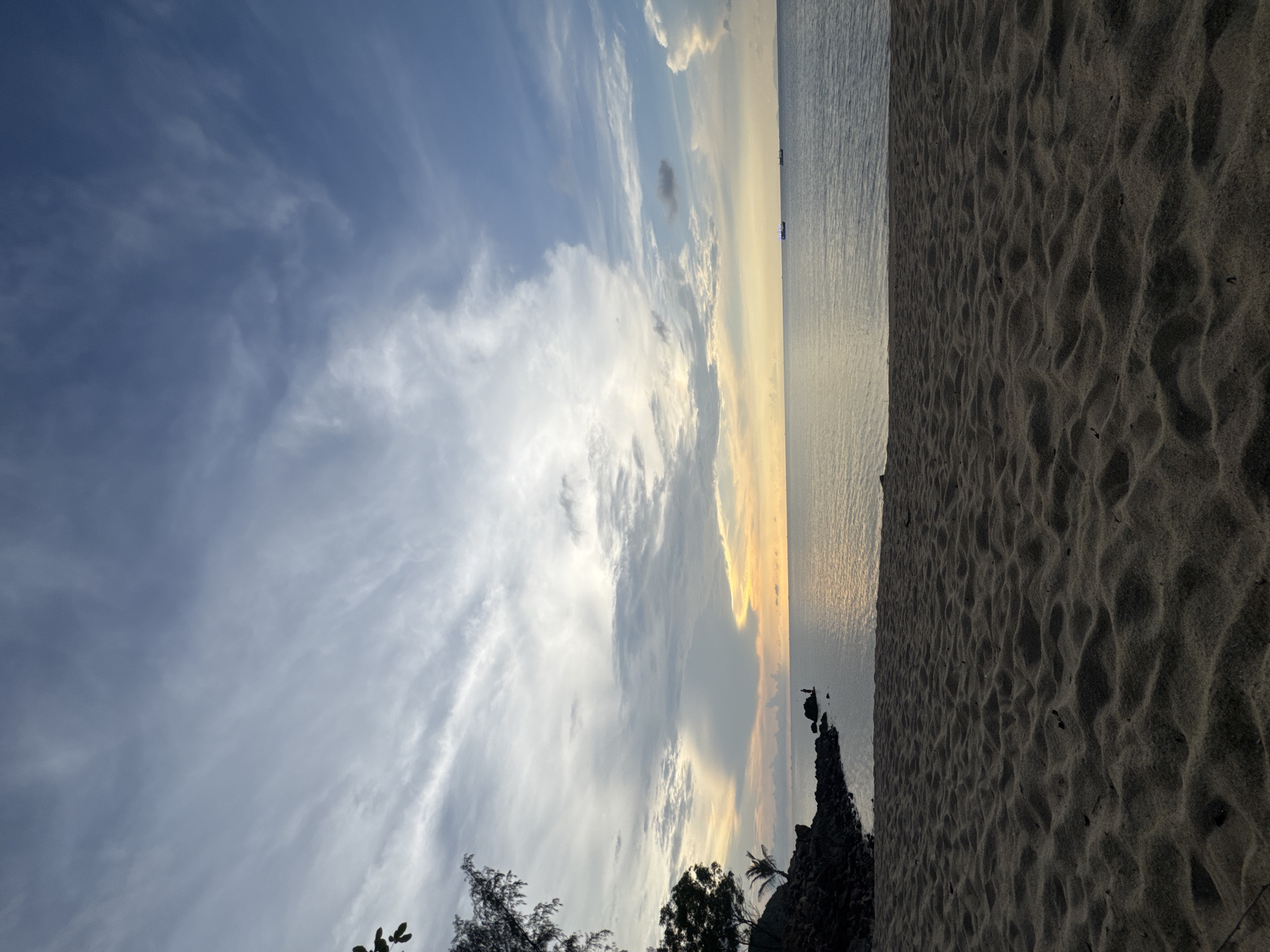 The image depicts a beach scene in Baan Wang Ta Kien, Thailand, oriented vertically. The sky occupies a significant portion of the frame, showcasing a mix of light blue and cloud formations, transitioning to hues of orange and yellow towards the horizon. The horizon line is relatively straight, separating the sky from the sea. The sea appears calm with a reflective surface mirroring the colors of the sky. A strip of sand occupies the right side of the image. Some vegetation is visible in the lower portion of the image along with rocks bordering the beach.