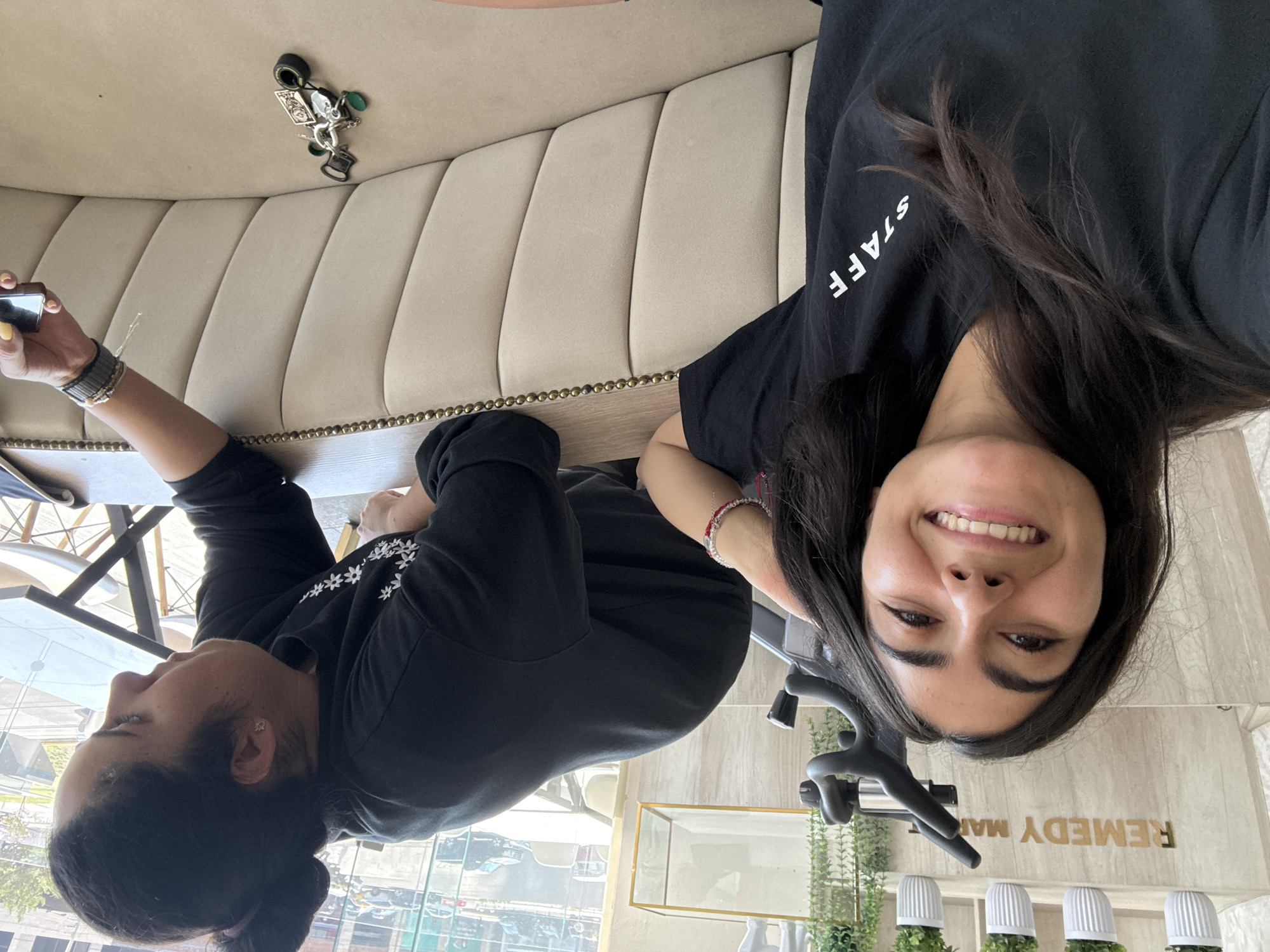 This image captures two women seated indoors, likely in a cafe or establishment, bathed in bright daylight. The scene suggests a relaxed atmosphere.
On the right, a woman with dark hair looks directly at the camera, smiling warmly. She is wearing a black t-shirt with "STAFF" printed in white on the left sleeve. A red string bracelet adorns her left wrist. Behind her, a gold sign on a light-colored wall displays the partial text "REMEDY MA," suggesting the business name, possibly "Remedy Market." Below the sign, several small green plants in white ribbed pots are arranged.
To her left, another woman, also with dark hair tied back, is engrossed in her phone, holding it in her right hand. She wears a black long-sleeved top featuring delicate white embroidered flowers on the sleeve and multiple bracelets on her left wrist.
Both women are seated at a wooden table with a distinctive studded trim, positioned alongside a comfortable, light beige, vertically tufted booth seat. A keyring with an assortment of unique charms, including a playing card, a top hat, and green-colored items, is visible resting on the booth next to the table.
Large windows behind the women offer a view of a sunny urban environment, with cars and buildings visible, suggesting a city setting consistent with Puebla, Mexico. The bright light streaming through the windows indicates it is daytime. The overall mood conveyed is pleasant and relaxed.