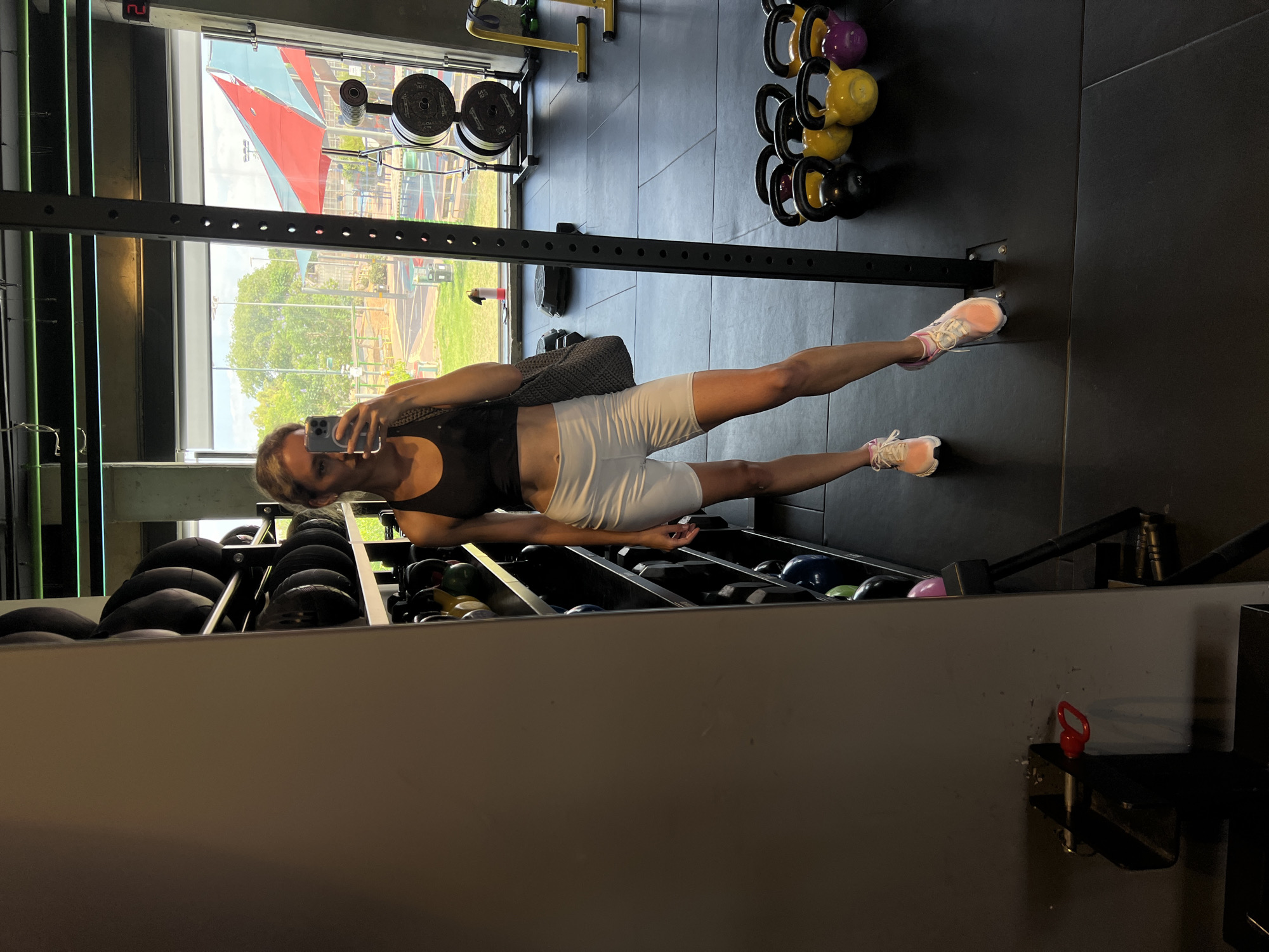 A woman is taking a mirror selfie in a gym. She is wearing a black sports bra and white biker shorts, with pink and white sneakers. She is holding a gray tote bag over her left shoulder. The gym has black rubber flooring, and there are racks of weights and kettlebells visible. Large windows look out onto an outdoor area with trees and what appears to be a playground. The scene is set in the city of Hod HaSharon, Israel.