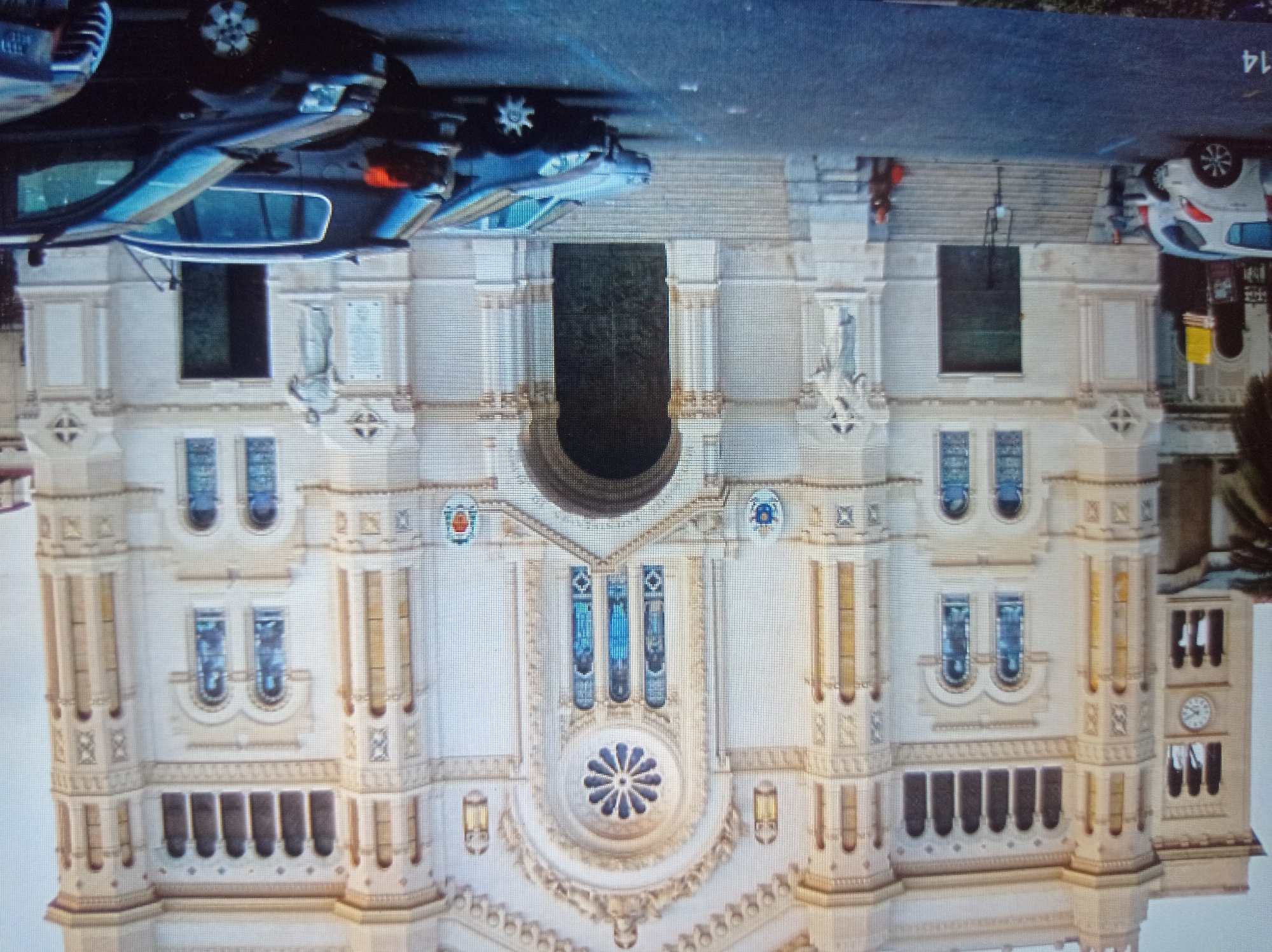 The image shows the facade of a large, ornate stone building, likely a church, with architectural details such as arched windows, decorative carvings, and a large central entrance. The building's facade is light-colored, possibly cream or white. In front of the building, there is a paved area that appears to be a parking lot. Several cars are parked in the lot, and two individuals are visible near the entrance stairs of the building. One person stands near the steps, and another sits on the steps on the right side of the image. The environment suggests an urban setting. The provided context indicates the location is Mukachevo, Ukraine, though this cannot be visually confirmed from the image itself.
