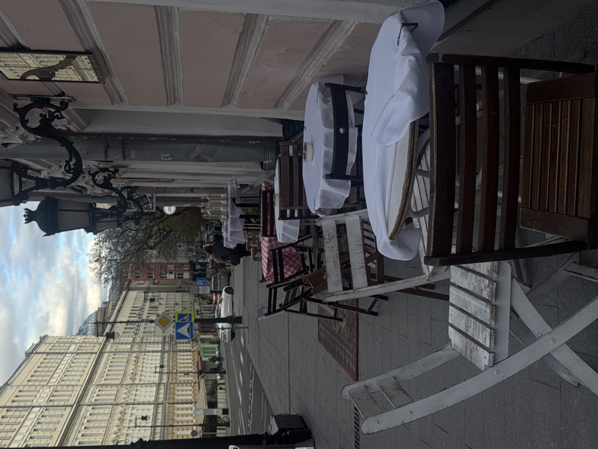 The image captures an outdoor dining area of a cafe or restaurant set up along a city street in Moscow, Russia. The scene is viewed from an angle looking down the street, with the cafe's tables and chairs occupying the right side of the frame.
Numerous tables, mostly covered with white tablecloths, are arranged on the sidewalk. Some chairs are light-colored or white, while others are dark wood. One table further down features a red and white checkered tablecloth. A patterned rug is laid out on the paved sidewalk beneath some of the tables. Ornate, dark metal street lamps with intricate scrollwork are visible along the building facade, one of which is partially reflected in a small mirror mounted high on the wall.
On the left side of the street, a grand, multi-story building with numerous windows and classical architectural details dominates the background. Further down, another building with a distinctive dome is visible. The street itself has parked cars, including a white sedan, and several traffic signs are present: a yellow diamond indicating a priority road, a blue square with a white arrow for straight ahead, and a blue 'P' sign for parking. The trees visible in the distance have bare branches, suggesting it is autumn, winter, or early spring.
The sky is overcast, indicating a cloudy daytime. Only one person is faintly visible far down the street among the tables. The cafe appears to be empty of customers, suggesting it might be closed, awaiting opening, or during off-peak hours. There is some faint text on a sign that appears to say "TAH" or "PAK" and "PO" or "DO", along with the clear "P" for parking.