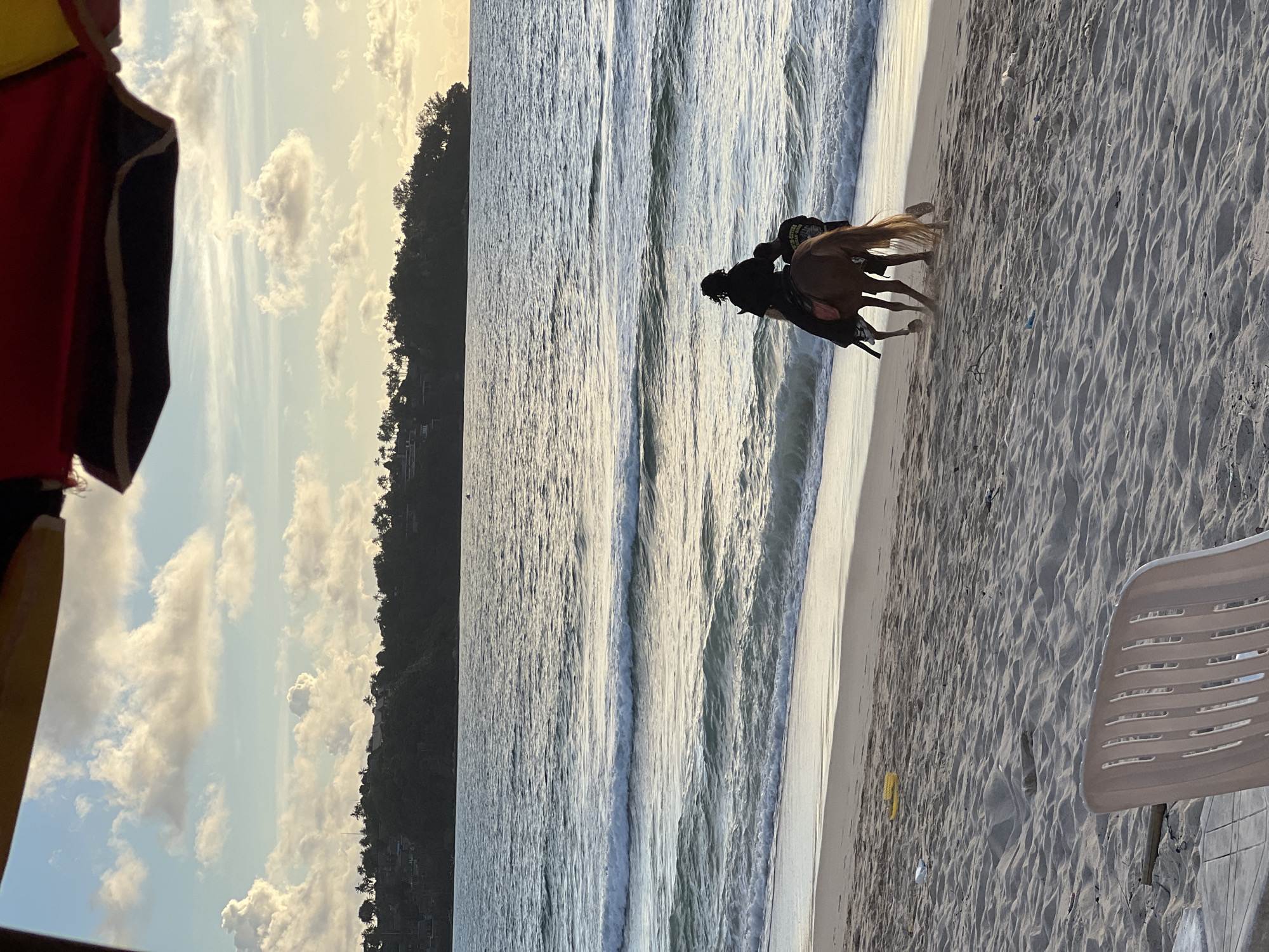 An aerial view captures a serene beach scene at sunset in Choeng Thale, Thailand. Two individuals are on horseback near the shoreline, with one person appearing to be riding while the other walks alongside. The horse is a reddish-brown color, and both individuals are dressed in dark clothing. The ocean waves gently roll onto the sandy beach, creating a peaceful atmosphere. In the background, a lush green hillside is visible, dotted with palm trees and homes, suggesting a tropical setting. The sky is a mix of soft clouds and fading sunlight, casting a warm glow over the entire landscape. In the foreground, a white plastic chair and a table are partially visible, indicating a seating area for beachgoers. The sand is scattered with some debris, including plastic waste.
