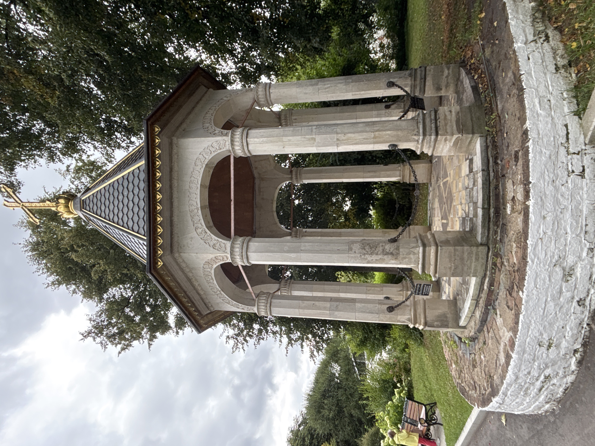The image shows a white, octagonal gazebo with a dark tiled roof and a golden cross on top. The gazebo is supported by eight stone pillars, each adorned with decorative carvings. Chains are suspended between the pillars, and a plaque is visible behind the chains. The ground inside the gazebo is paved with patterned tiles. The structure is surrounded by a low, circular brick wall, painted white, with some visible damage. Behind the gazebo, there is lush green foliage and trees under a cloudy sky. To the left of the gazebo, a park bench sits on a grassy area, and a person wearing a yellow jacket and red pants is seated on the bench, facing the gazebo. The location is identified as the Savvino-Storozhevsky Monastery in Zvenigorod, Russia.