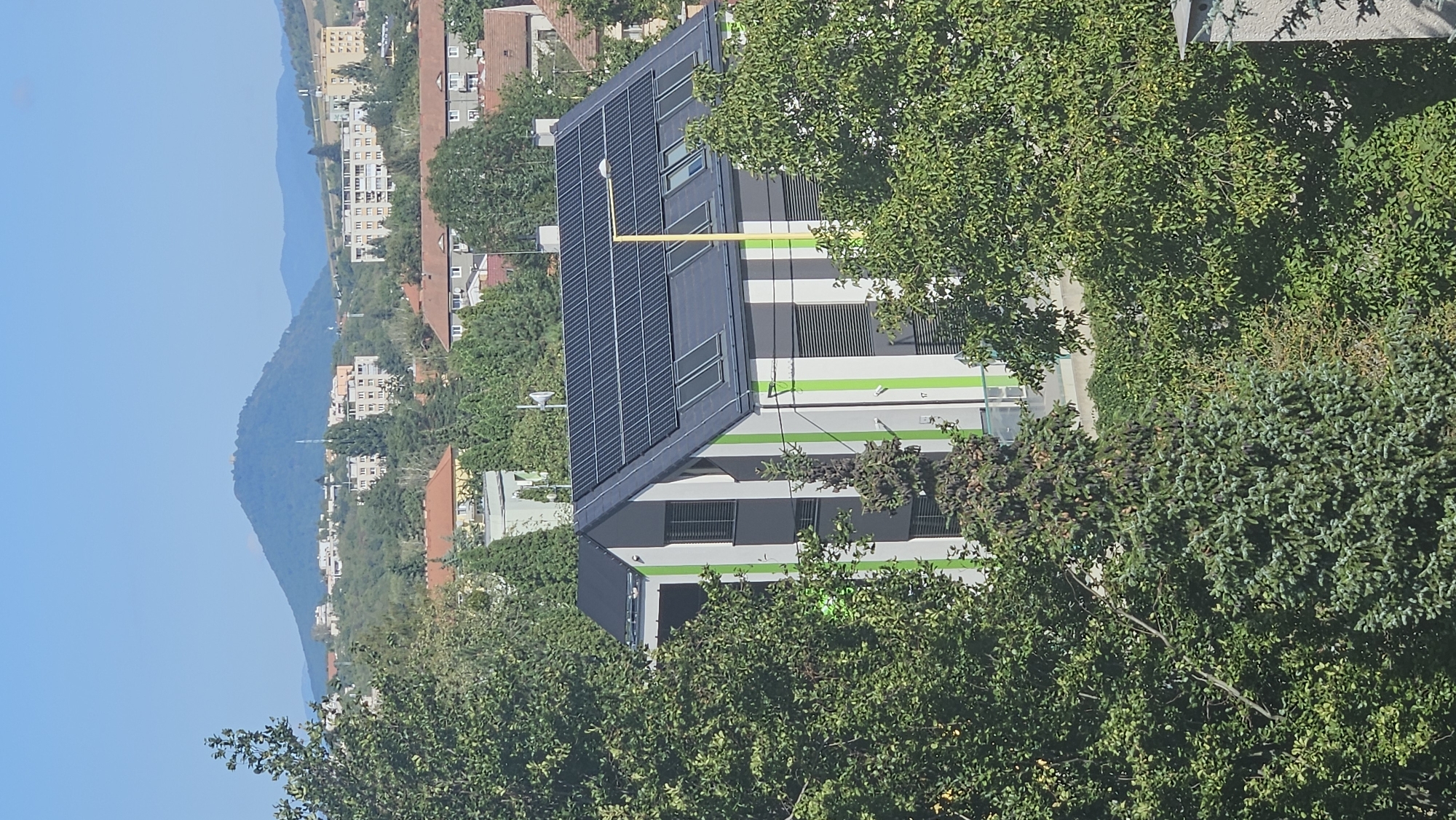 A vertical, medium shot captures a modern building with a striped facade and a solar-paneled roof, nestled amongst lush green trees in the city of Prešov, Slovakia. The building, painted with alternating vertical stripes of white, grey, and lime green, features numerous windows and skylights. A tall, bright yellow pole, possibly a streetlamp, stands in front of the building. In the background, a prominent, round, tree-covered mountain with a small structure on its peak dominates the skyline. Below the mountain, residential buildings and more trees are visible, contributing to the urban and natural landscape. The sky is a clear, bright blue. The image is taken from a slightly elevated vantage point, looking down on the scene. No people are visible in the frame.
