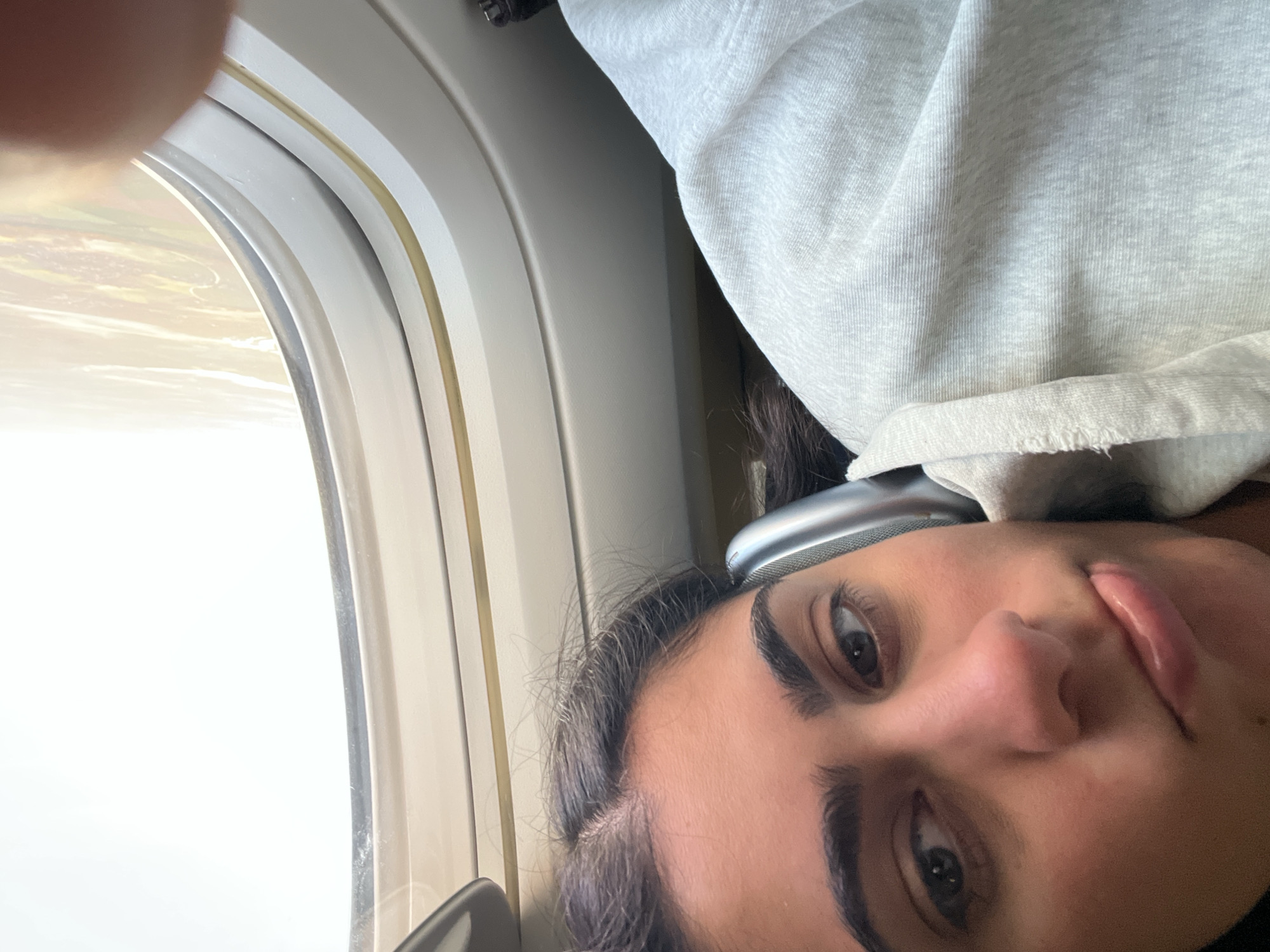 A close-up selfie of a person with dark hair, brown eyes, and a slightly parted mouth, potentially female, taken inside a plane near the window, with the window frame and a blurred view of the outside visible. The person is wearing headphones. The lighting is bright. The location, Meaux, France, cannot be determined from this image alone.