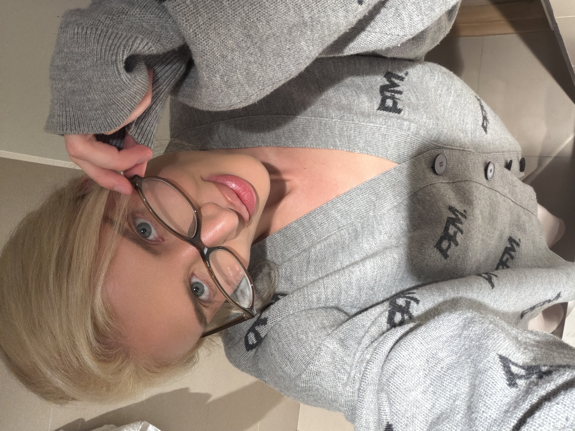 The image depicts a blonde female in an indoor setting. The subject, positioned centrally, is observed from a slightly elevated angle. She has blonde hair pulled back, light blue eyes, and is wearing a light grey V-neck cardigan with three visible dark grey buttons down the front. The cardigan is patterned with multiple instances of black "PM." letters. Her left hand is raised, holding brown-framed eyeglasses, with the lenses positioned over her eyes. Her lips appear full and glossy. The individual is looking directly at the viewer with a slight smile. The background consists of plain, light-colored walls, possibly beige or off-white, with a darker horizontal surface visible below, suggesting a countertop or similar fixture. The lighting is uniform. This scene is reported to be located in the city of Belgrade, Serbia.