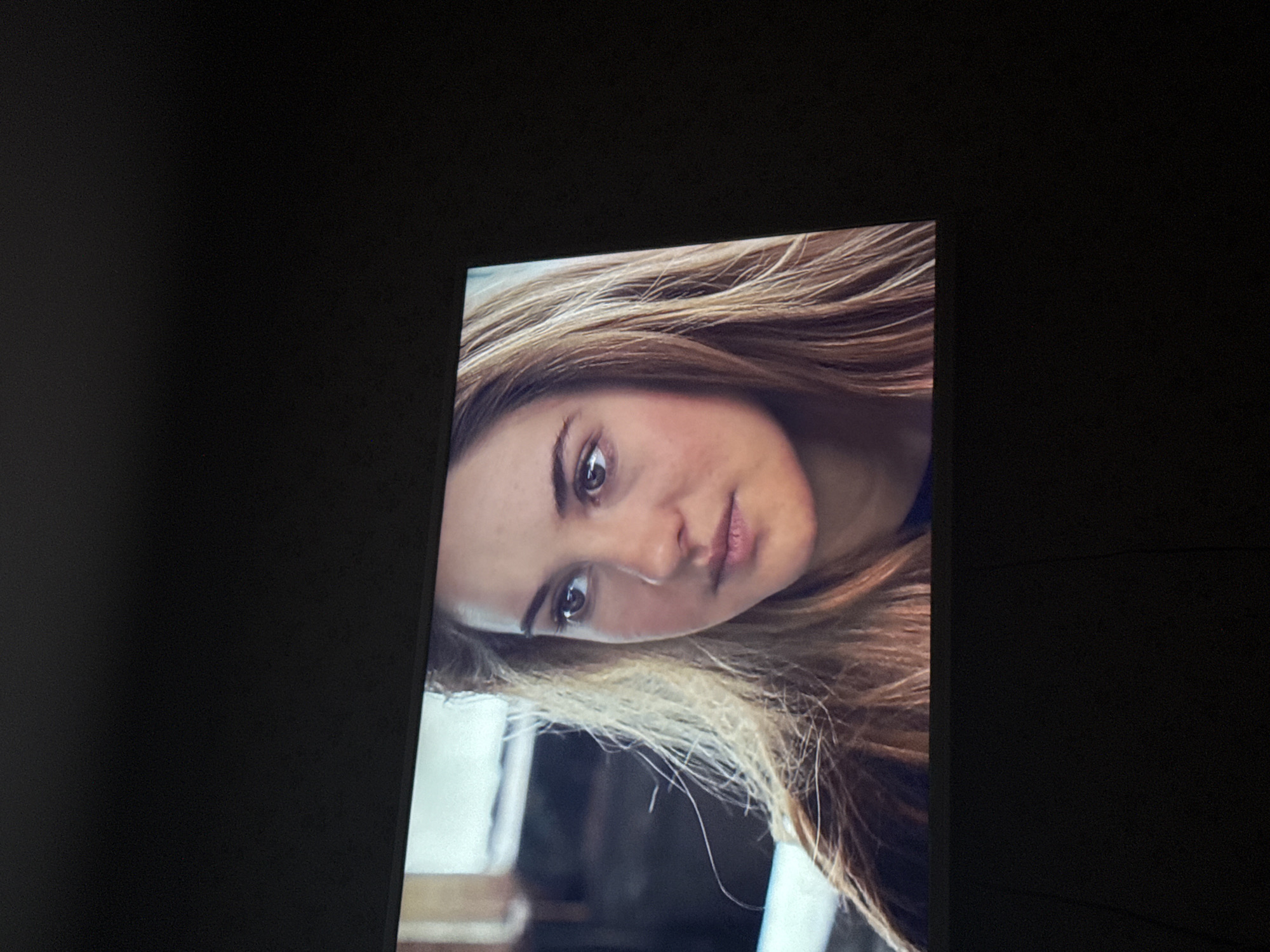 A television screen displays a portrait of a fair-skinned woman with blonde hair and brown eyes. She has a neutral expression and is looking slightly downwards. The background visible on the screen appears to be an indoor scene, though the details are somewhat blurry. The television screen is positioned in front of a solid, dark background. The image was taken indoors. The location is unknown and not within Mytishchi, Russia.