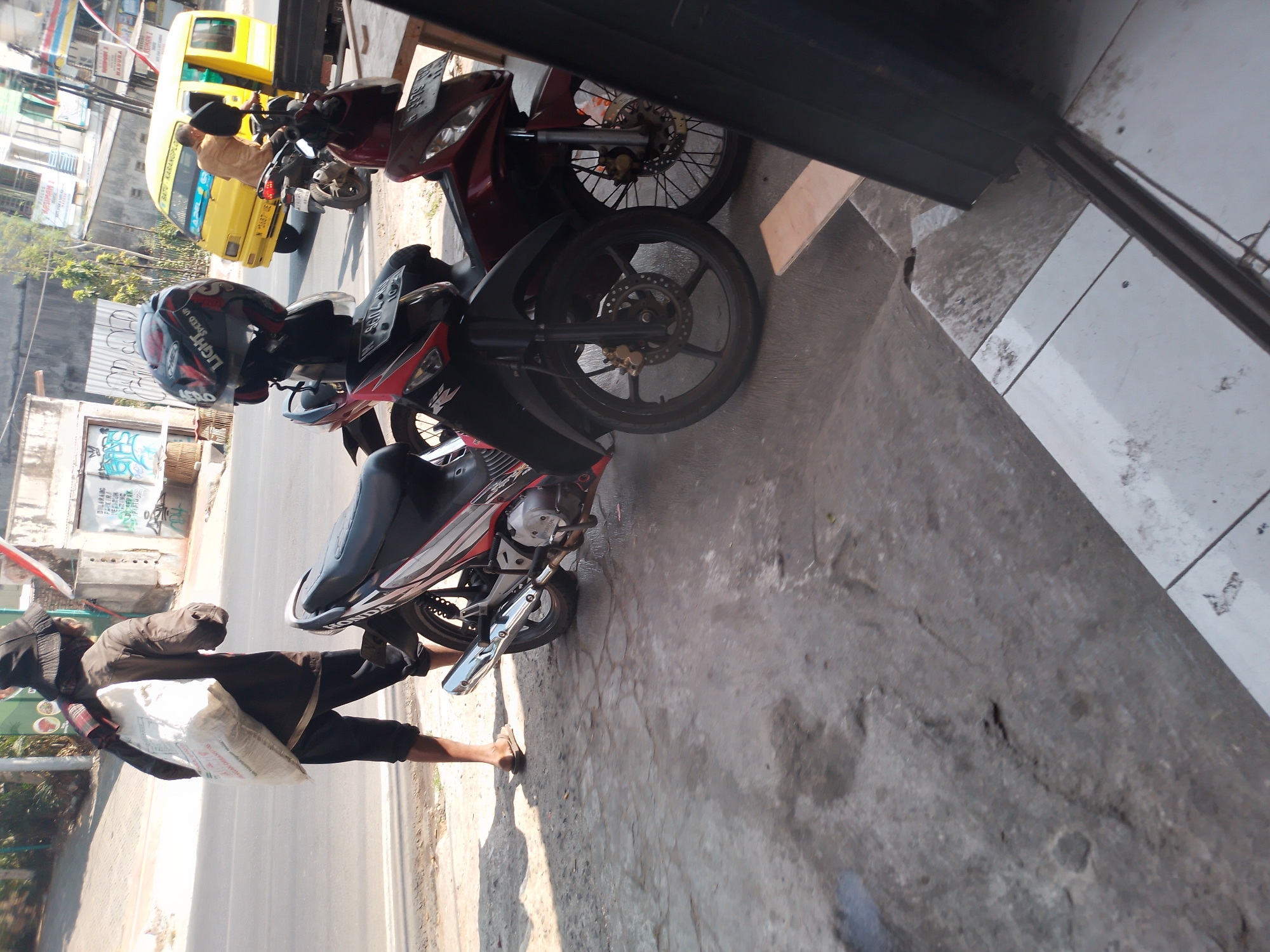 A person in a hat and dark clothing walks on a cracked concrete sidewalk, carrying a large white sack over their left shoulder. The person is walking away from the camera, towards a street. Two motorcycles are parked on the sidewalk in front of the person, facing towards the street. The motorcycle closest to the camera is dark red, and the one behind it is black and red. A yellow van is driving on the street in the background, and other buildings are visible. The scene appears to be in Batu, Indonesia, based on the presence of a yellow van commonly used as public transportation there.