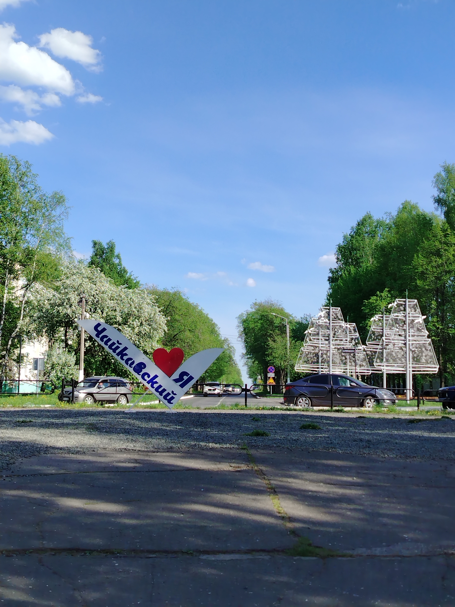 The image depicts an entrance to the city of Chaikovsky, Russia. In the foreground, the gray concrete ground is cracked and uneven. A sign bearing the name of the city, "Чайковский Я" (Chaikovsky I), written in blue Cyrillic letters, with a red heart symbol, leans towards the left. A silver SUV is visible behind the sign. In the background, a street lined with green trees leads toward the city center. On the right side of the street, there are abstract, geometric metal tree-like structures. Dark cars are parked or moving along the road. The sky is blue with scattered white clouds.
