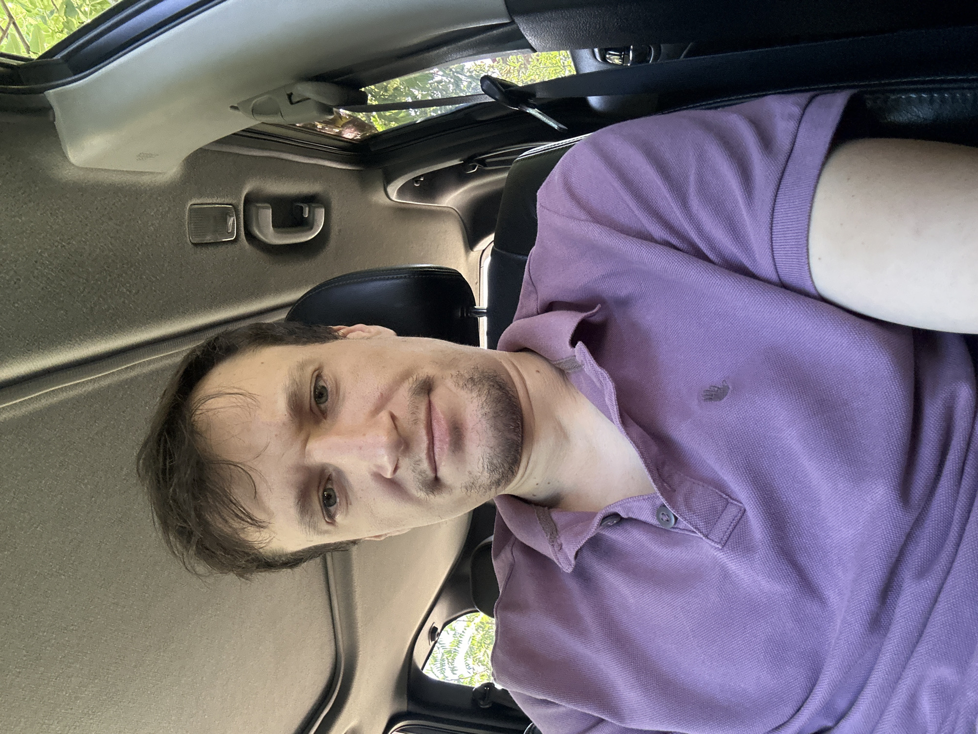 A man is sitting in the driver's seat of a car, likely in Makiv, Ukraine. He is wearing a purple polo shirt and has short, dark brown hair and a mustache. His facial features are visible, and he appears to be looking directly at the camera. The car's interior is visible, including a sunroof, a headrest, and a seatbelt. Sunlight filters in from the windows, illuminating the interior and the surrounding trees. The scene is brightly lit, suggesting it is daytime.