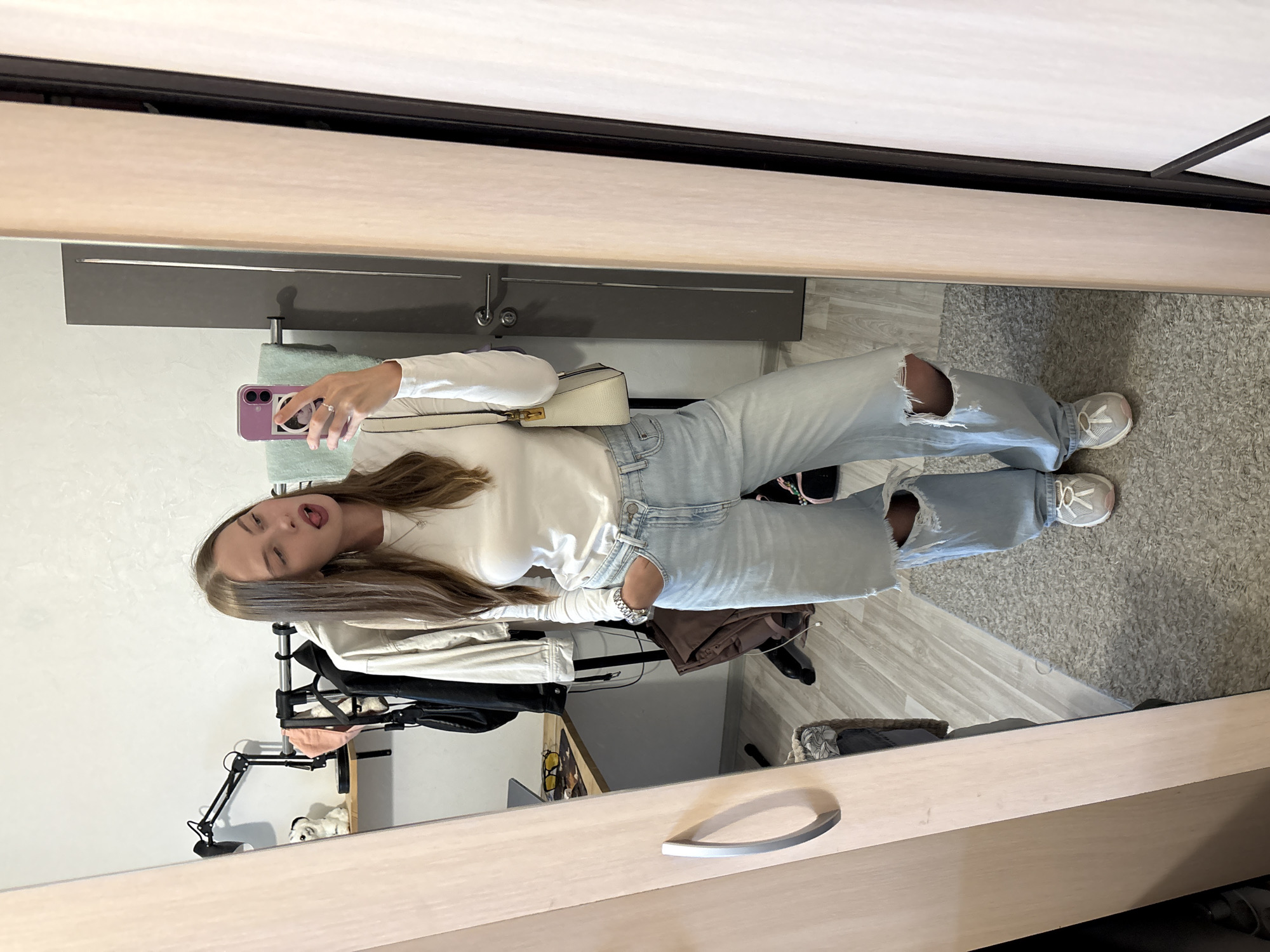 A young woman with long brown hair stands in front of a full-length mirror, taking a selfie. She is wearing a white long-sleeved shirt, light-wash ripped jeans, and white sneakers with pink accents. She is holding a light-colored handbag with a strap over her shoulder and a purple phone in her right hand. Her left hand is in her pocket, and she is sticking her tongue out towards the camera. The mirror reflects a room with a grey carpet, a desk with a laptop and yellow glasses, a clothing rack with several garments, and a grey door in the background. The woman is positioned in what appears to be a bedroom or dressing area within an apartment. The setting is consistent with a residential interior in Perm, Russia.