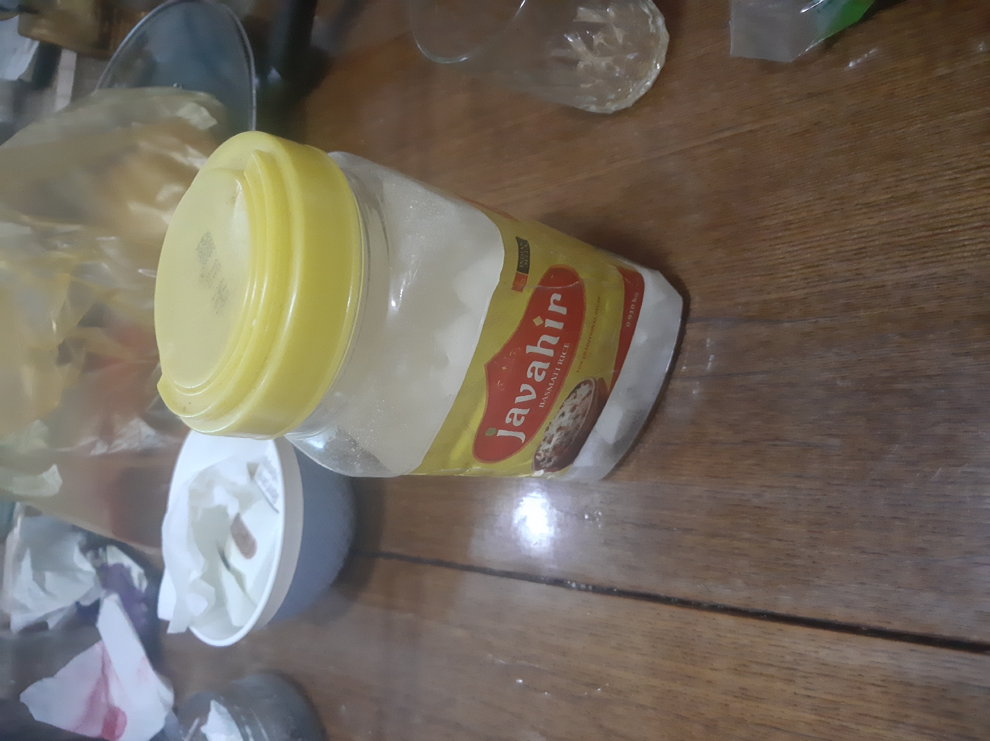 A clear jar of basmati rice with a yellow lid sits on a wooden table. The jar has a label that reads "Javahir" and "BASMATI RICE". A crumpled white tissue is in a blue and white bowl to the left of the jar. A glass is to the right of the jar. The image appears to be taken indoors in a residential setting. There is no indication of location in Sabunchu, Azerbaijan.