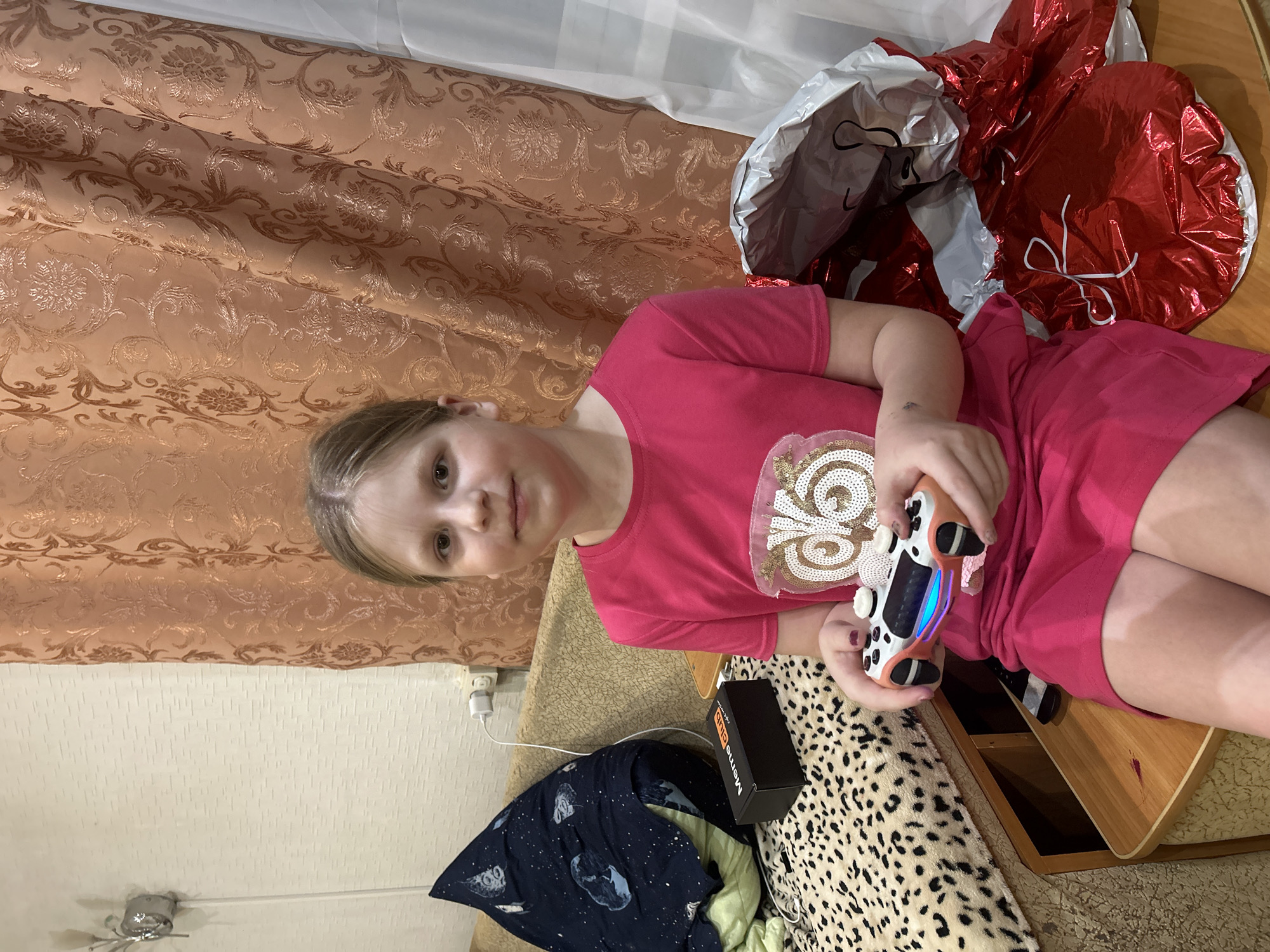 A young girl sits on a wooden surface, likely a desk or shelf, holding a video game controller. She wears a bright pink shirt with a sparkly, circular decoration on the front, and a matching pink dress. Her light brown hair is pulled back, and she looks directly at the camera with a neutral expression.
Behind her are a curtain with an ornate floral pattern, as well as several crumpled, possibly deflated, red and silver balloons. Beside her, resting on a leopard-print blanket, is a black box with the words "Meme Club" printed on it. A navy blue blanket patterned with celestial illustrations is also visible. A charger is plugged into the outlet.
Based on the information provided, the location is Фоки, Russia.