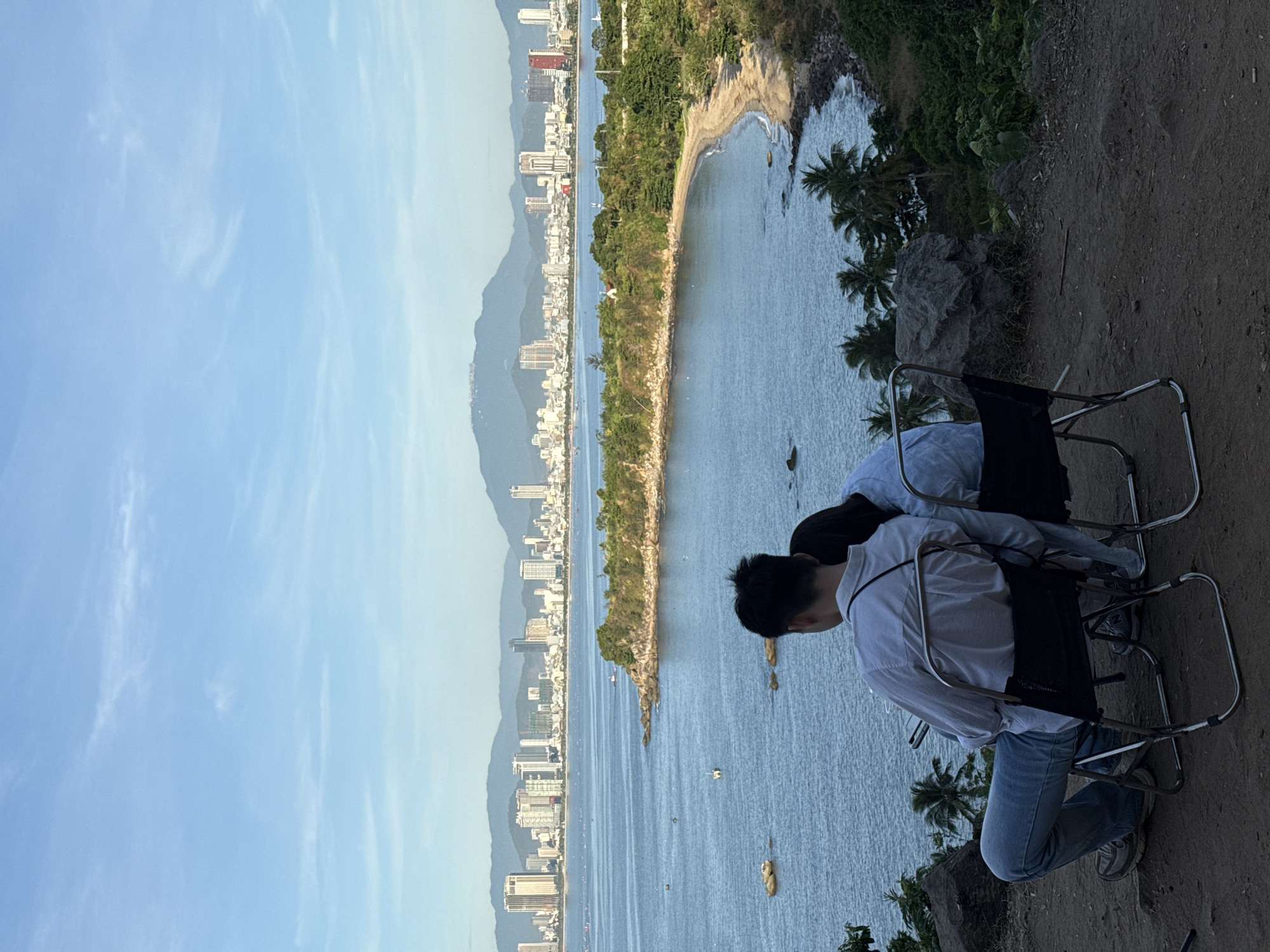 A man and a woman, viewed from behind, sit on folding chairs on a hilltop overlooking a bay. The man is wearing a light blue collared shirt and jeans, holding a phone in his left hand. The woman is wearing a light blue shirt. The bay is calm, with several small boats visible. In the distance, a curved beach leads to a treed peninsula. Beyond the peninsula, a cityscape with numerous high-rise buildings lines the coast, backed by hazy mountains. The sky is clear with wispy clouds. The scene is set in Sơn Trà Peninsula, Đà Nẵng, Vietnam, likely at a viewpoint offering panoramic vistas of the city and the East Sea.