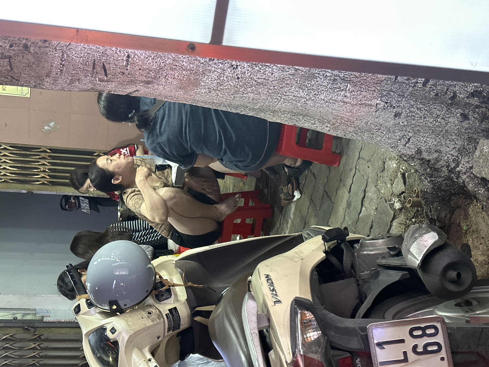 The scene appears to be a street view in Đà Nẵng, Vietnam, showing several individuals sitting on red plastic stools next to a parked Honda Vision scooter. The scooter has a light gray helmet on the seat, and its license plate reads "17-L1 68." The people are sitting closely together, suggesting a casual and potentially social atmosphere. The surroundings include a building with a roll-up door, a concrete wall, and street debris.