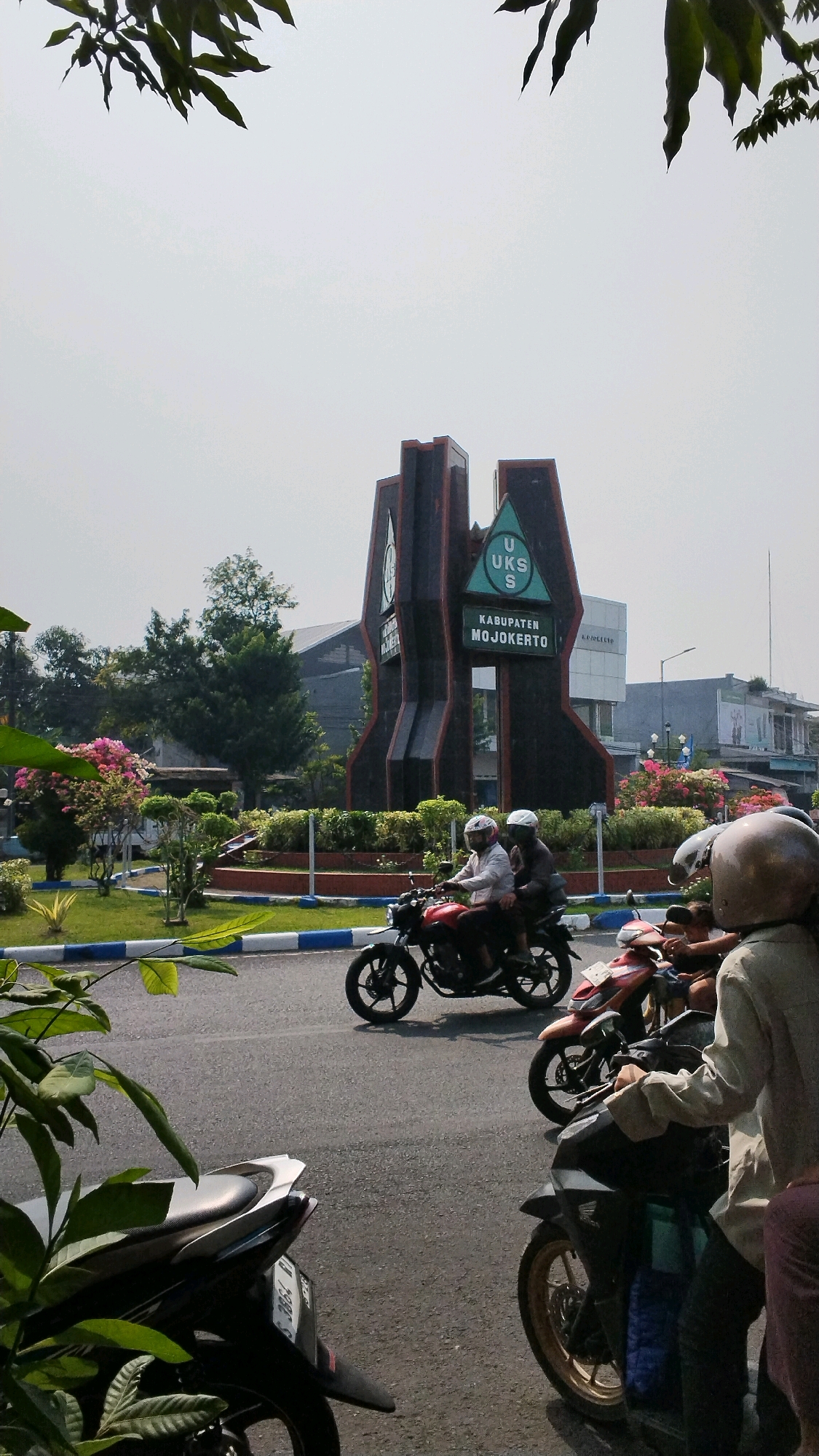 In the city of Banjaragung, Indonesia, a monument stands tall, its dark, angular structure accented with green and white signage that reads "UKS S KABUPATEN MOJOKERTO." The monument is surrounded by a well-maintained garden with blooming pink flowers and manicured shrubs. A road runs in front of the monument, where several motorcycles are present. Two individuals are riding on a red motorcycle, both wearing helmets. Another motorcycle, partially visible, is to the right. A third motorcycle, a dark blue scooter, is parked in the foreground on the right, with a person wearing a light-colored shirt and a helmet standing next to it. The sky above is a pale, hazy white, suggesting a bright, possibly hot, day. Green leaves from an overhanging tree frame the top of the image, casting a slight shadow and adding a natural element to the urban scene.