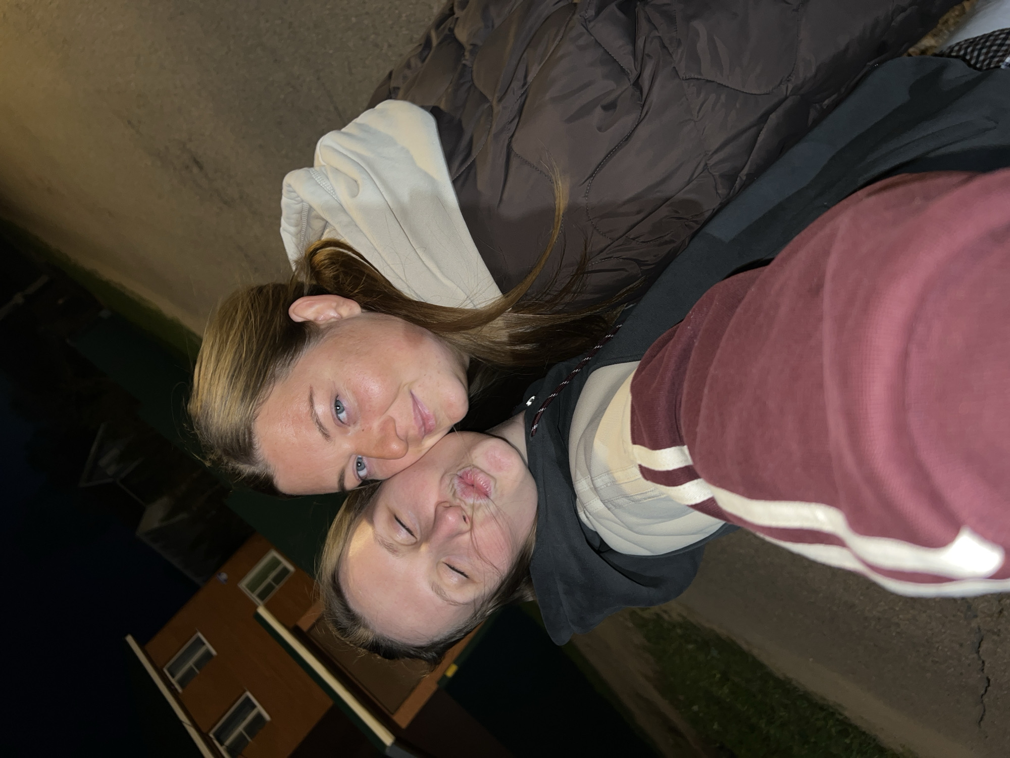 Two young women are posing closely together for a photograph. The woman on the left has fair skin, light brown hair, and blue eyes. She is looking at the camera with a slight smile. The woman on the right is making a kissing face with her eyes closed. Both are wearing casual clothing. One is in a brown puffer jacket with a beige hoodie layered underneath. The other is wearing a burgundy and white striped hoodie over a black jacket and beige hoodie. In the background, a two-story brick building with lit windows is visible, suggesting it is nighttime. The ground appears to be a street or sidewalk. This scene is located in the city of Раздолье, Russia.