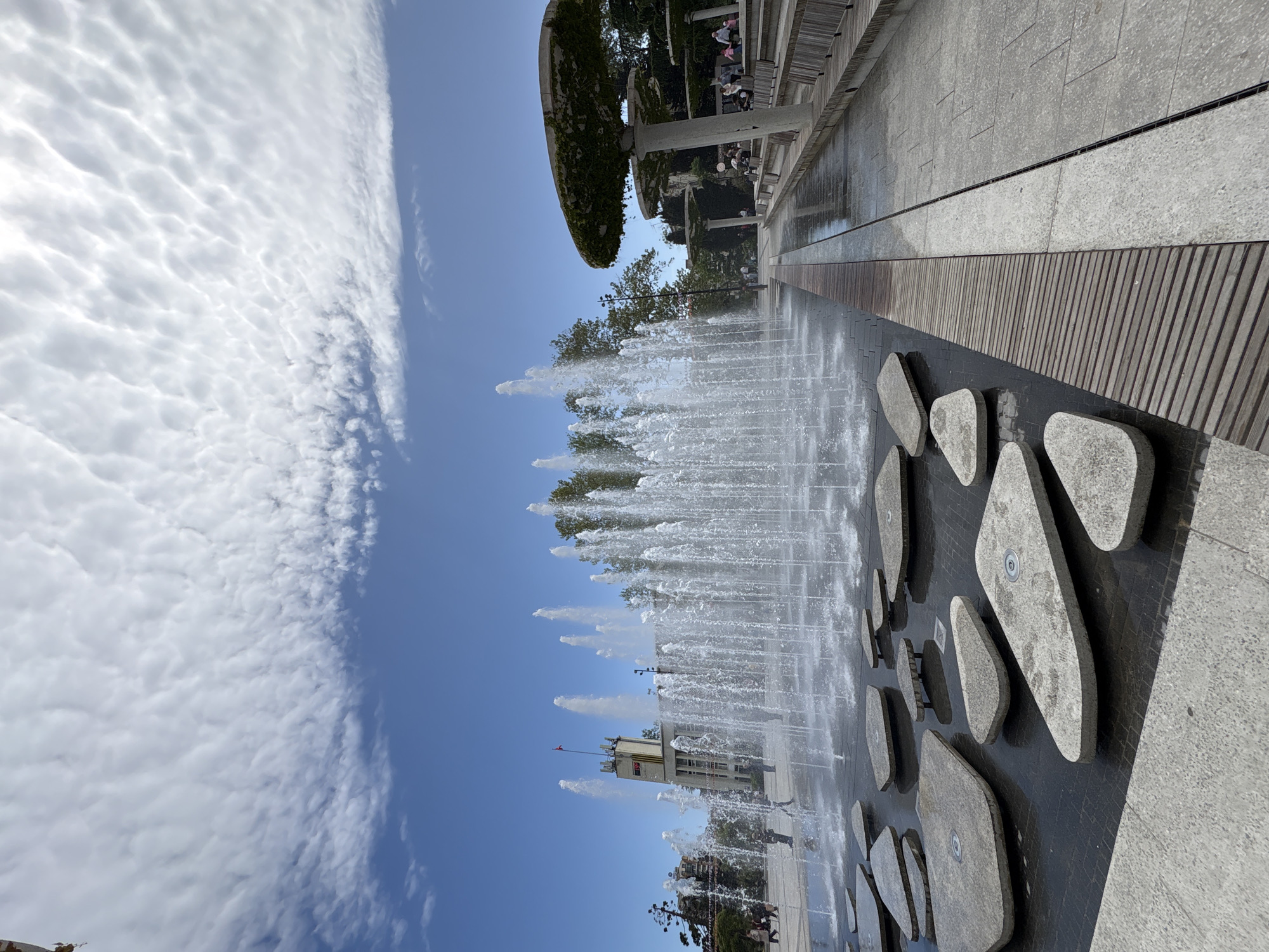 A vertical outdoor shot depicts a fountain with multiple jets of water spraying upwards. In the background is a building with a red flag, presumably the city hall in Yalta, Ukraine. Above the fountain is a clear blue sky partially filled with textured white clouds. The foreground consists of a dark tiled surface with embedded stone-like shapes. People are faintly visible in the background, enjoying the space. The location is the city of Yalta, Ukraine.