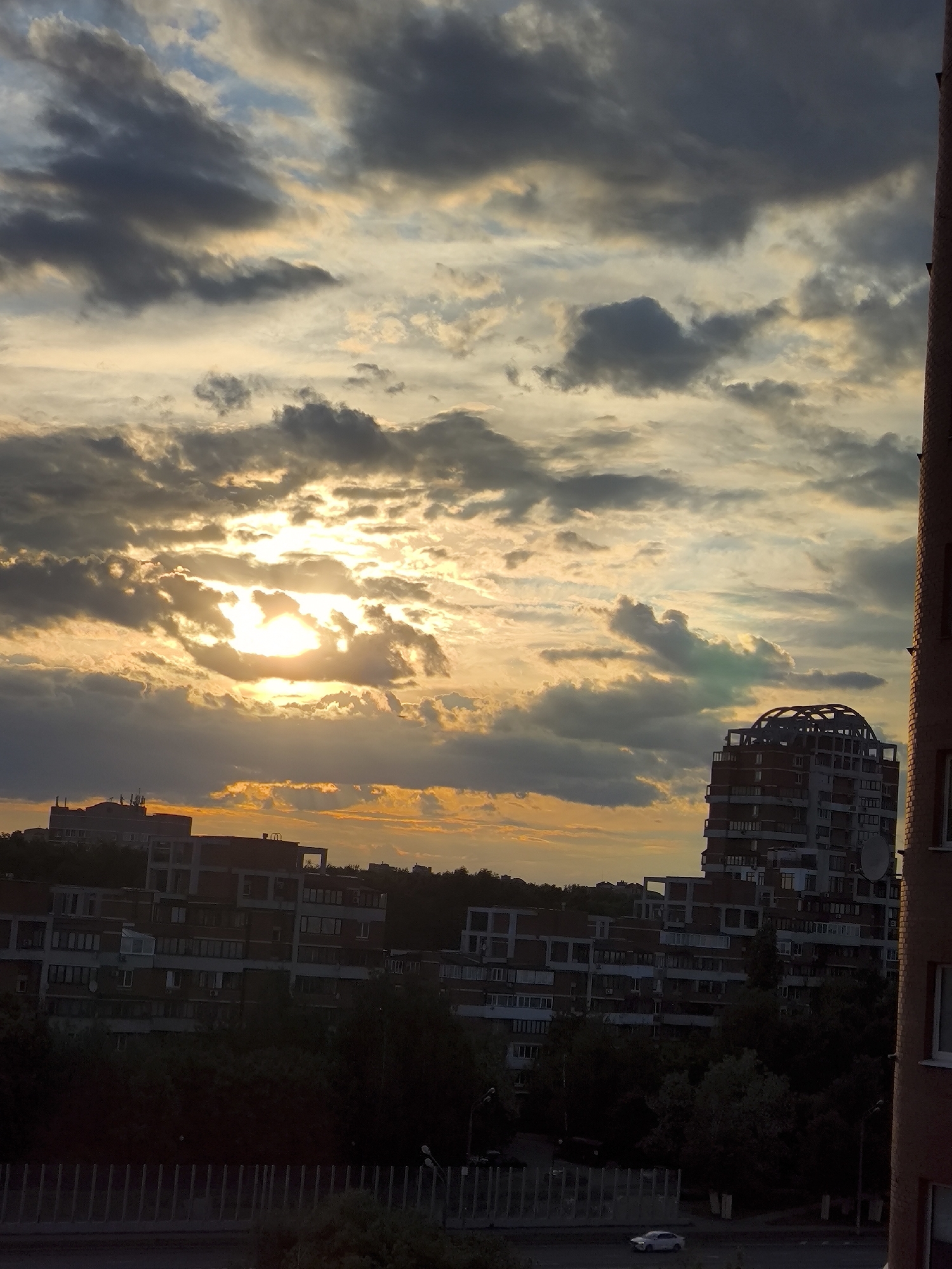 The image displays an urban or suburban landscape viewed from an elevated perspective, likely at sunset. The sky dominates the upper portion of the frame, filled with a dramatic arrangement of clouds. The sun, a bright, golden orb, is partially obscured by dark, textured clouds, creating vibrant sunrays and illuminating the sky with warm orange and yellow hues. Patches of pale blue sky are visible between the cloud formations.
Below the horizon, several multi-story residential buildings are visible, largely silhouetted against the bright sky. On the left, a block of buildings with numerous windows and balconies stands. To the right, a distinctive taller building features a unique rounded structure on its roof and a large satellite dish on its facade. A brick building, partially cropped, frames the extreme right side of the image.
Dark, dense foliage and trees are interspersed among the buildings, suggesting green spaces. A street runs horizontally across the lower part of the image, where a single white car is present, appearing to drive or be parked. A dark fence or barrier stretches across the immediate foreground. The scene evokes a tranquil urban evening. There is no visible text in the image.