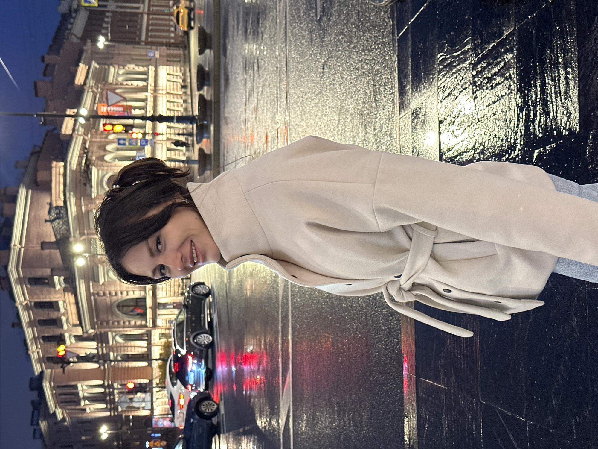 A woman is standing on a wet sidewalk in Saint Petersburg, Russia. She is wearing a light-colored coat with a belt tied around her waist. In the background, a historic building is illuminated with street lights and there are a number of vehicles on the street. The image is rotated 90 degrees clockwise.