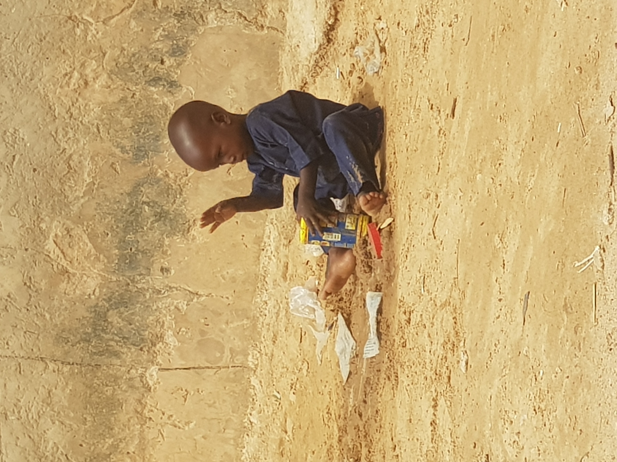 A young child sits barefoot on sandy ground strewn with litter in Dutsinma B, Nigeria. He wears a dark blue outfit and plays with a box. A wall is visible in the background.