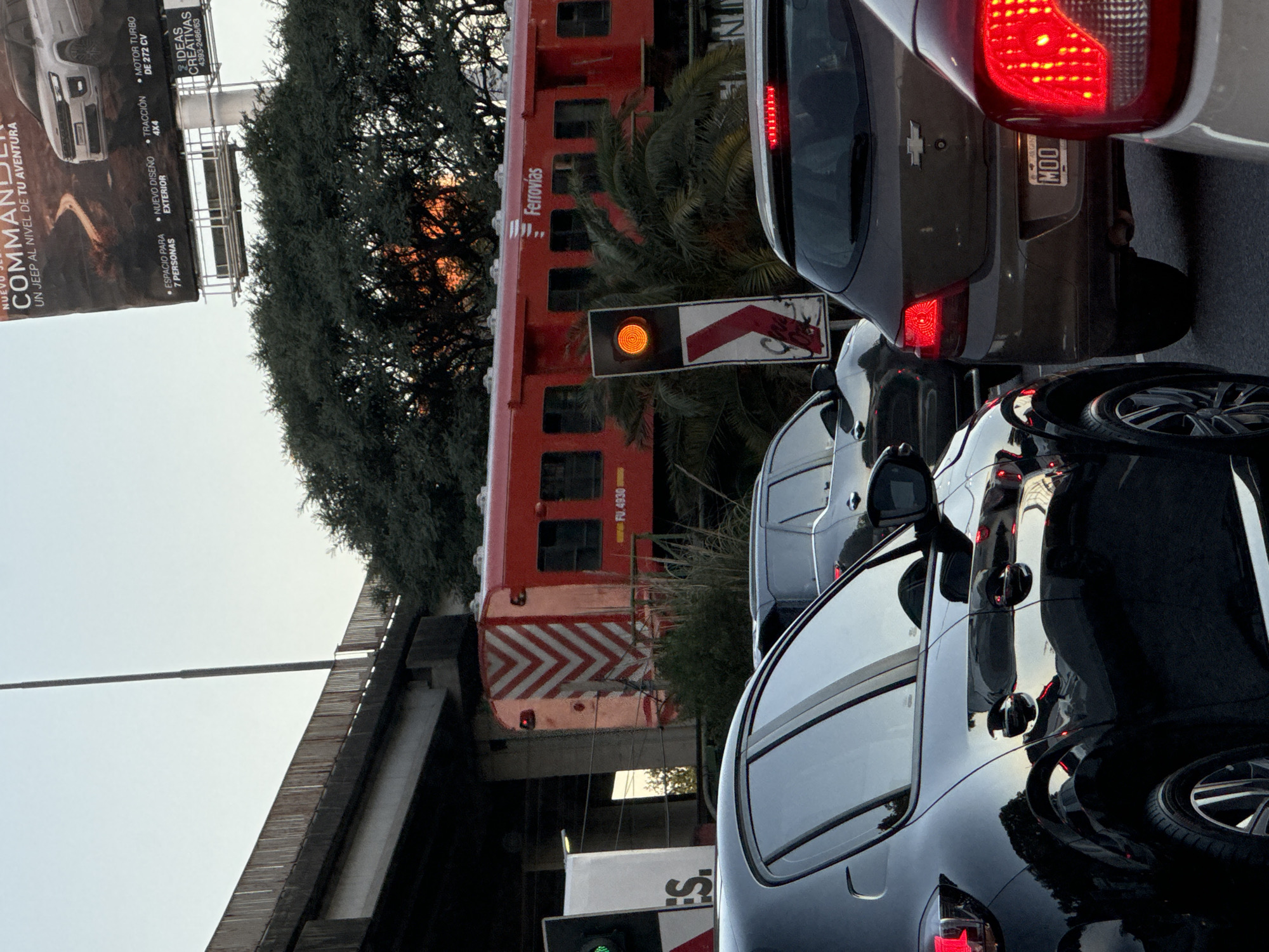 Here is a detailed description of the image:
The image shows a traffic scene in Buenos Aires, Argentina, with a train passing overhead. Several cars are stopped in traffic. In the foreground, there is a black car, with a dark gray car behind it with the license plate "MOO". The red tail lights of both vehicles are visible. The license plate indicates that it is a vehicle from Argentina. A red train labeled "Ferrovias" is passing over a bridge. Visible signage on the train identifies it as "FU. 4930". A traffic light with an orange light is visible next to the train. A billboard advertising a Jeep Commander is also visible on the left of the image. The environment includes trees, infrastructure (bridge), and other elements of a city street.