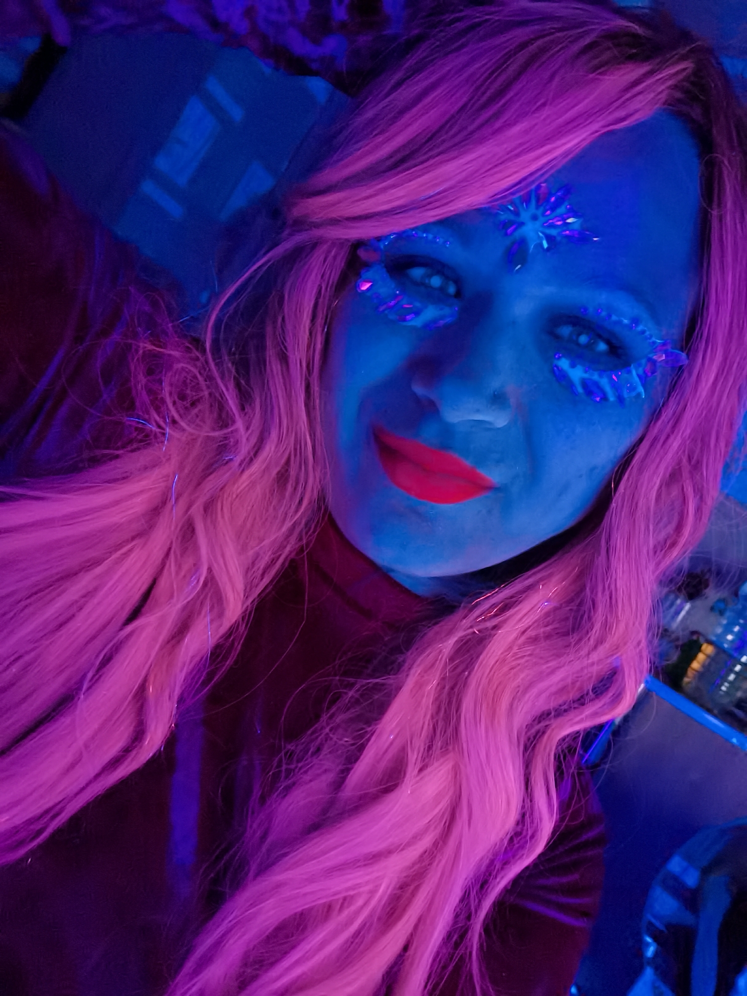 A person with pink hair and blue skin stands illuminated by a blue light, possibly in a club or party setting. Their face is adorned with blue-toned jewel-like accents around their eyes and forehead. They are wearing a dark-colored garment, and their lips are colored bright red. The location is unknown.