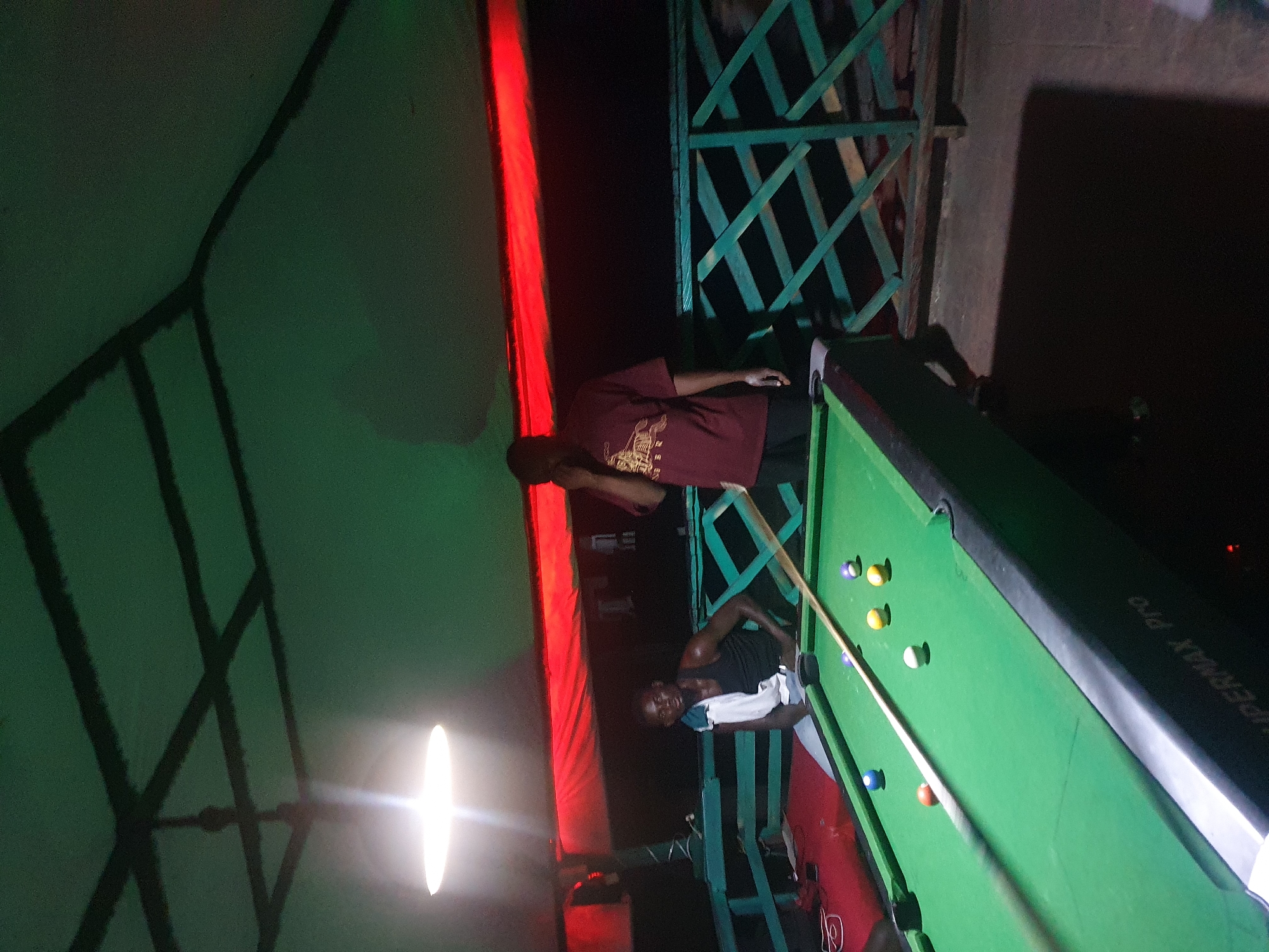 The image depicts an indoor or covered outdoor scene, possibly a bar or recreational area, featuring a pool table. Two men are present. One man, wearing a black tank top and grey pants, is seated at the edge of the pool table, with a white towel draped over his left shoulder. The second man, wearing a maroon t-shirt with a graphic design and dark pants, stands near the table, holding a mobile phone to his ear. The pool table is green, with several colored balls and a cue stick visible. The environment is dimly lit by an overhead light, and a red fabric is draped across the background, suggesting a themed or decorative element. The overall impression is a casual gathering focused on playing pool. The location is specified as Lokoja, Nigeria, though the visual elements do not provide specific landmarks to confirm this.