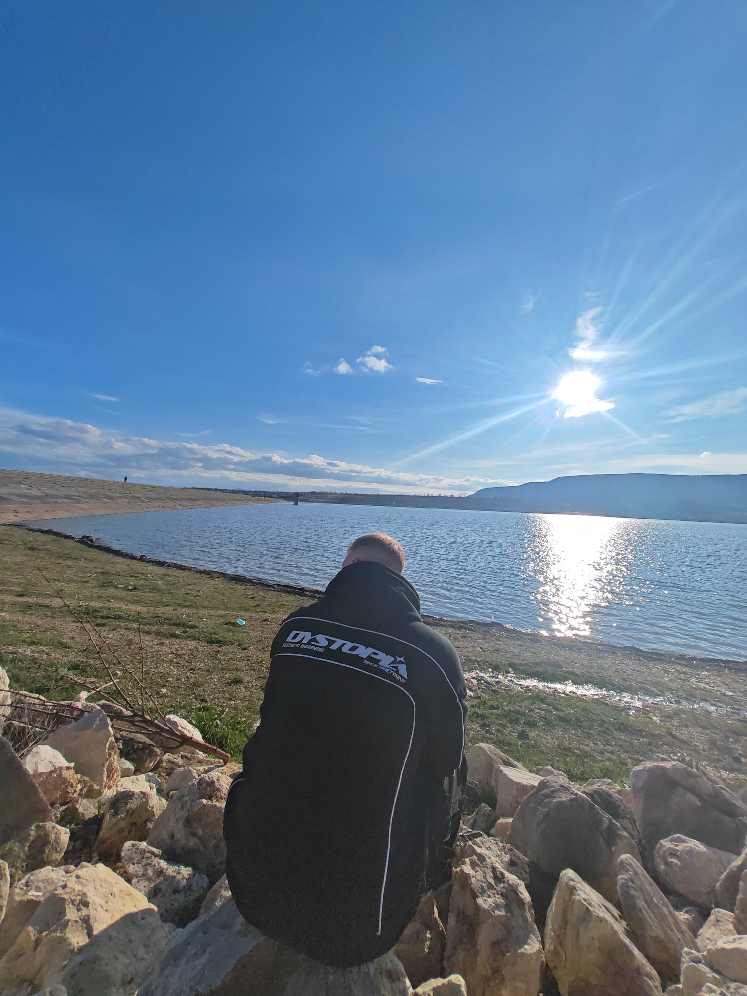 A person sits on a pile of rocks facing a large body of water on a sunny day. The individual is wearing a black jacket with white lettering on the back reading "DYSTOPIA AESTHETIC AWARENESS SINCE NINETEEN NINETYNINE". The water reflects the sunlight, creating a shimmering effect. In the background, there are distant hills and a partially cloudy blue sky. The scene is likely at a reservoir or lake near Shumen, Bulgaria.