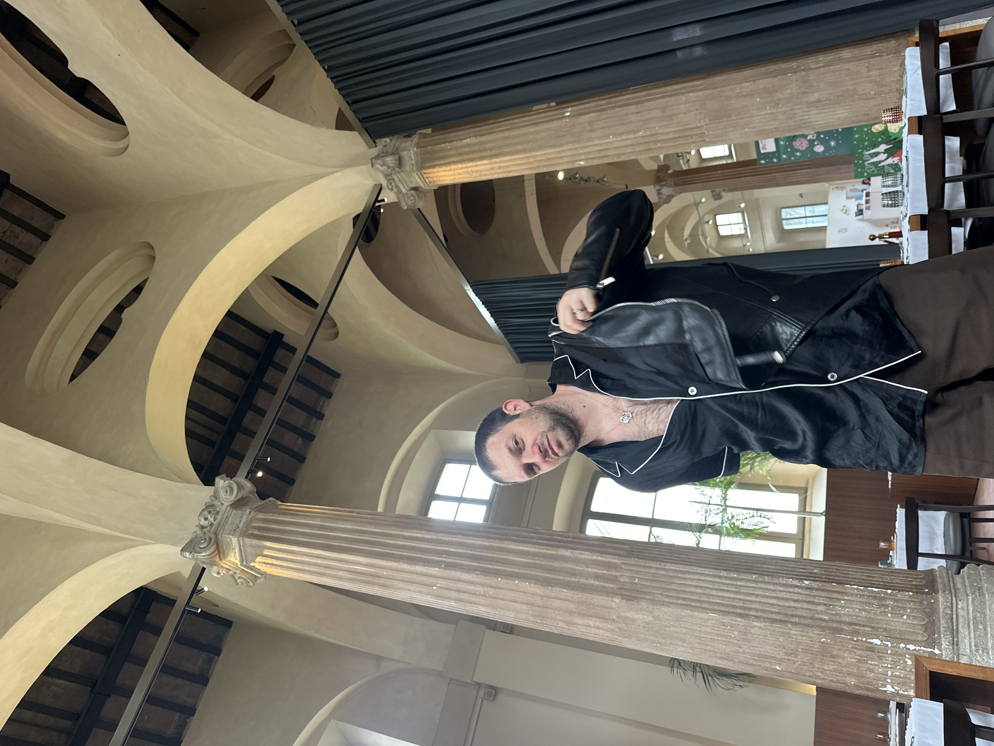 The image depicts an adult male posing in what appears to be a high-end restaurant or event space located within a historic building in Milan, Italy. Specifically, the architectural style, featuring fluted columns with ornate Corinthian capitals, multiple arched vaults, high ceilings, and tall windows, strongly suggests the interior of **Giacomo Bistrot** at Via Pasquale Sottocorno, 6, Milan.
The subject, a male with short dark hair and stubble, is positioned slightly off-center to the right, looking directly at the camera with a neutral expression. He wears a black satin-like shirt with white piping, partially unbuttoned to reveal chest hair and a silver-colored pendant necklace. In his right hand, extended to his side, he holds a black leather jacket with a zipper.
The environment is characterized by its grand, classical architecture. Several massive, light-colored fluted columns with decorative Corinthian capitals support an intricate system of arched vaults that form the high ceiling. Some sections of the ceiling reveal darker, possibly wooden, structural beams above the plastered vaults. Natural light enters through tall, rectangular windows set into arched wall openings in the background. Modern track lighting fixtures are mounted along the ceiling beams. Dark, heavy curtains are drawn along one side of the room, near another column. The space is set up with multiple tables, each covered with a white tablecloth and featuring dark chairs. Table settings include glassware and what appears to be small decorative elements. A large green plant is visible near one of the windows. The scene is well-lit, combining natural light from the windows with ambient artificial lighting.