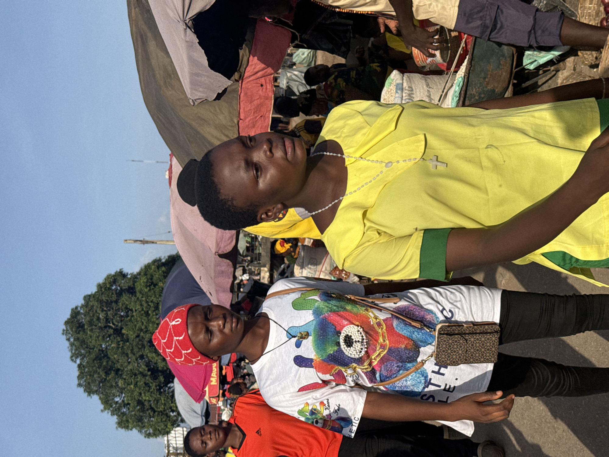 The image depicts a lively outdoor market scene, likely in Onitsha, Nigeria, under a clear, bright sky, suggesting it's daytime. The primary subjects are several young individuals, seemingly engaged in commerce or simply present in the bustling environment. One person, wearing a bright yellow shirt and a cross necklace, looks upwards with a contemplative expression. Another, sporting a red headscarf and a white t-shirt with a colorful graphic print, also appears to be looking away, possibly at a vendor or a passerby. A third person in an orange shirt is partially visible in the foreground, looking directly at the camera. The background reveals a dense collection of market stalls, indicated by various fabrics, bags of goods, and what appear to be fruits or vegetables. The overall atmosphere is one of activity and social interaction typical of a Nigerian market. The image is taken from a slightly tilted perspective, adding to the candid feel of the scene.