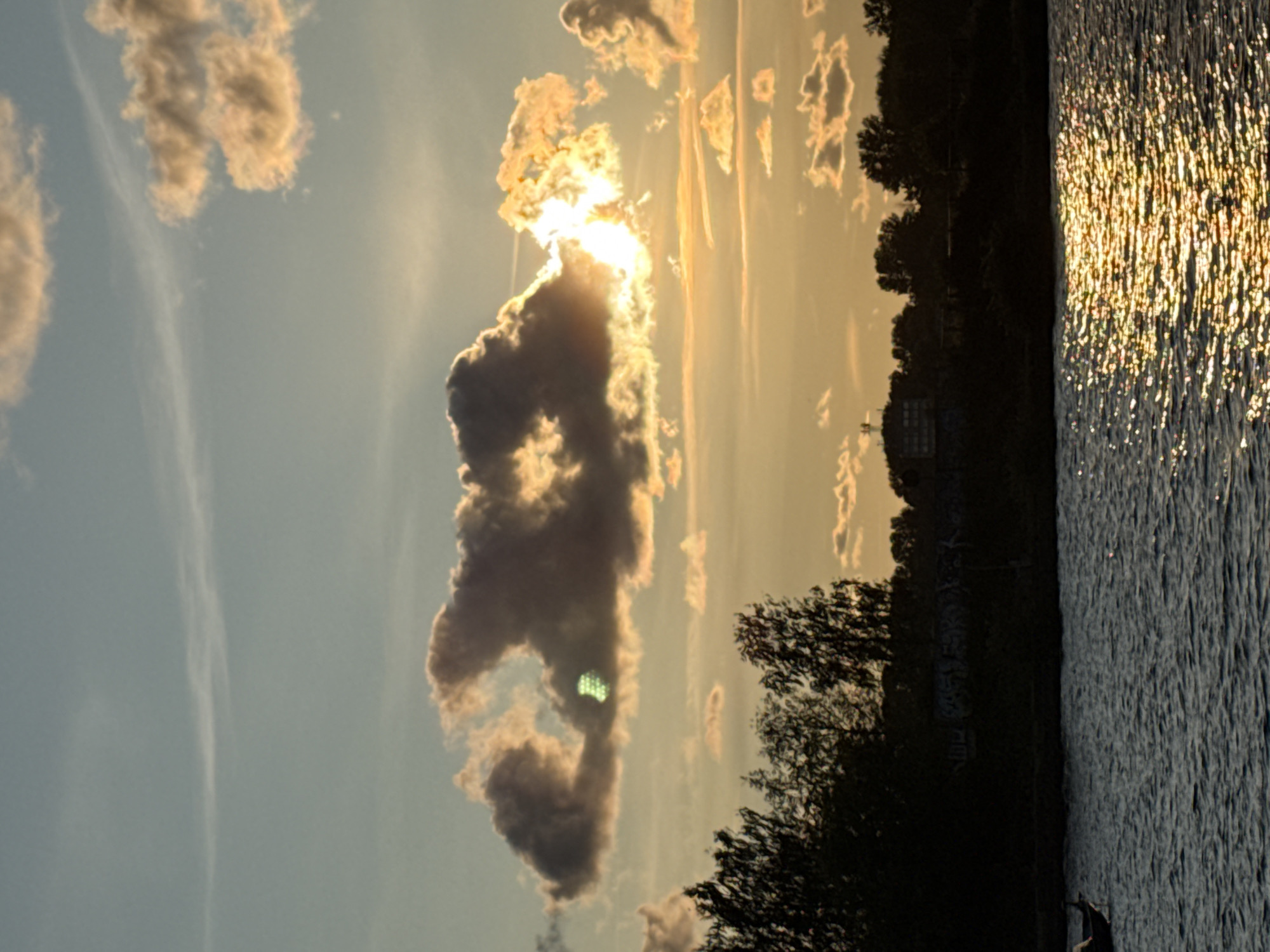 This image depicts a sunset over a body of water, likely the Daugava River, in Riga, Latvia. The sky is a gradient of soft blues and oranges, with scattered clouds tinged by the setting sun. A prominent dark cloud formation sits in the upper center, partially obscuring the sun, which emits a bright, hazy glow. A distinct green, pixelated light source is visible within this cloud. The foreground features rippling water reflecting the warm light of the sunset. Along the far bank, a silhouetted treeline runs across the horizon, with a few structures visible, including what appears to be a building or complex on the left side of the frame. The overall scene is tranquil, with a boat visible in the lower left quadrant, its silhouette dark against the water.