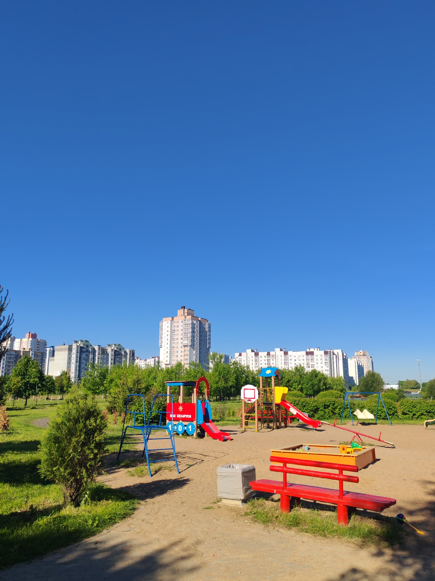 The image captures a playground in an urban setting. A clear blue sky dominates the upper part of the frame. The playground contains various structures, including a blue and red train-shaped play structure with a slide, a red bench, and a yellow sandbox. Other playground elements include a swing set, a seesaw, and a climbing structure with a basketball hoop. In the background, there are residential buildings surrounded by green trees, suggesting an urban park environment. The ground surface of the playground is sandy. The image appears to be taken during the day under bright sunlight.