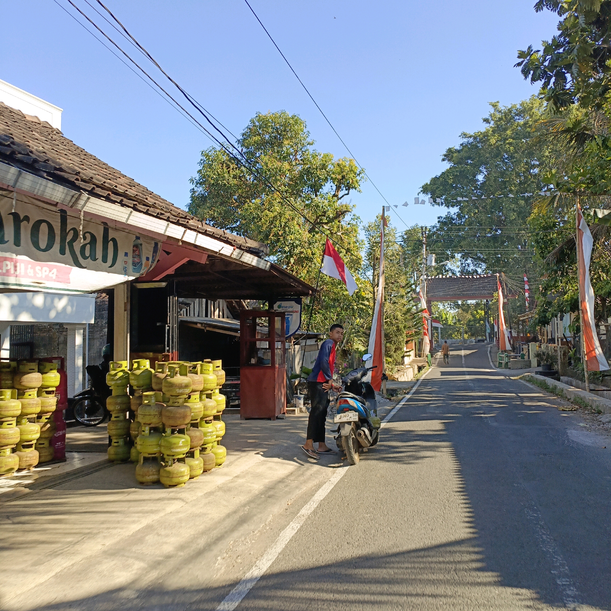 The image depicts a street scene in Salatiga, Indonesia, under a clear blue sky. In the foreground, a large stack of green gas cylinders is piled on the left side of the road, next to a building with a tiled roof and a banner that reads "Marokah". A young man wearing a blue and red shirt and dark pants stands next to a motorcycle on the right side of the road, facing the camera. The motorcycle is blue and has a license plate that reads "6303 SD". The road is paved and appears to be a street in a residential or commercial area, with trees and flags lining the sides. In the background, there is an arched structure over the road and more trees and buildings. The overall impression is a sunny day in a bustling Indonesian town.