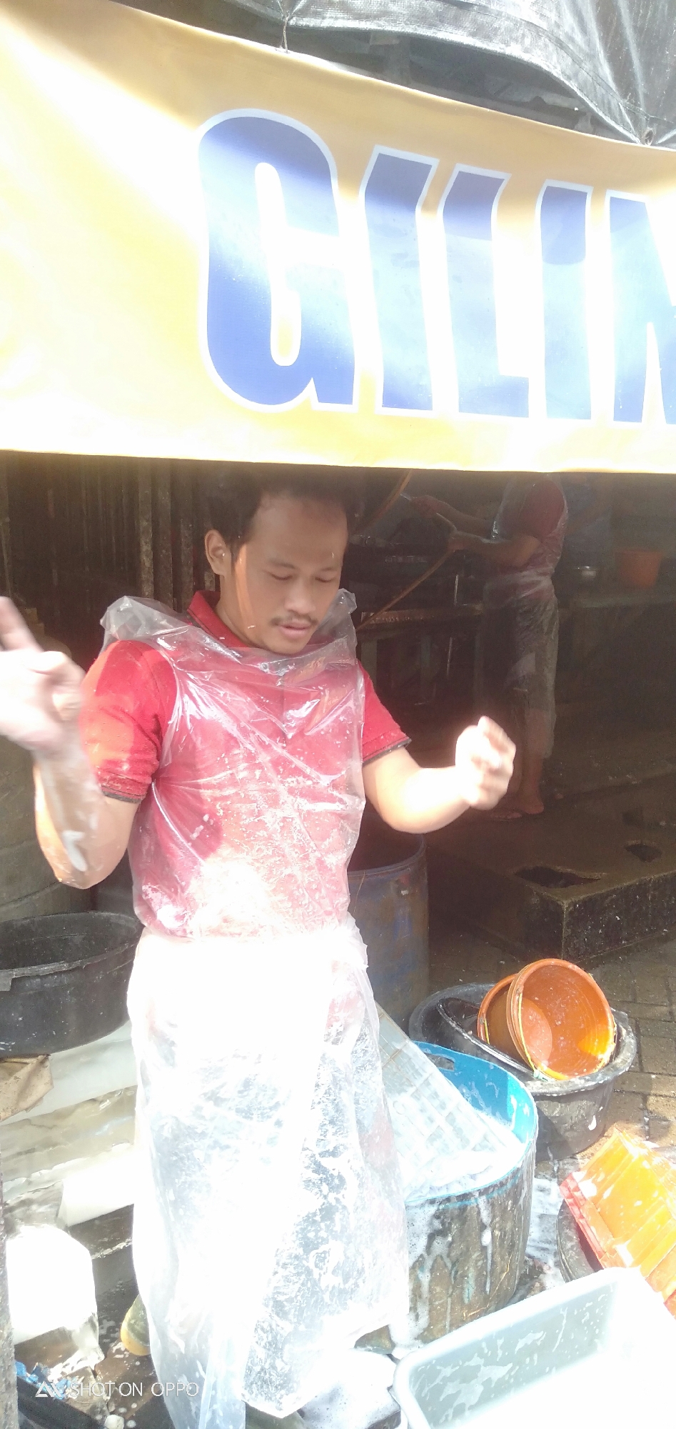 A man in a red shirt and plastic apron is standing in front of a yellow banner with blue lettering that appears to say "CILIN". He has his arms outstretched, and his hands are blurred with motion. Behind him, another person can be seen working at a stall, and several buckets are visible, some containing water or soapy suds. The scene appears to be an outdoor food stall or market, possibly in South Jakarta, Indonesia, given the text on the image. The lighting suggests it is daytime, with sunlight casting shadows. The overall impression is one of a busy, functional workspace.