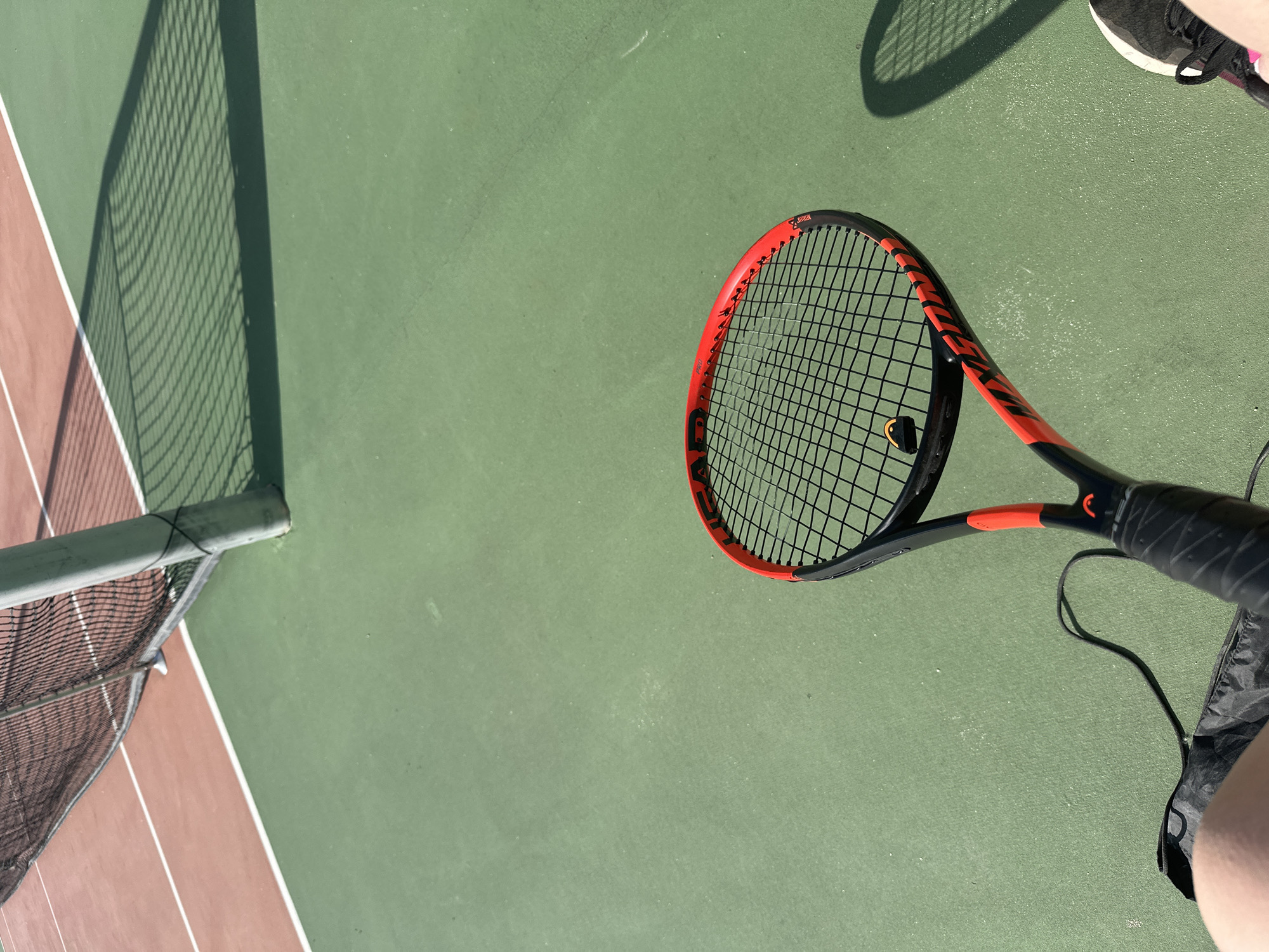 The image shows a tennis court, likely outdoors, with a tennis racket lying on the green surface. A net and post are visible on the left side, adjacent to a reddish-brown area, suggesting a border or another court. Part of a person's leg and shoe can be seen in the upper right corner. A black object is visible on the tennis racket.
There is insufficient information in the image to determine the location in the city of Sofia, Bulgaria.