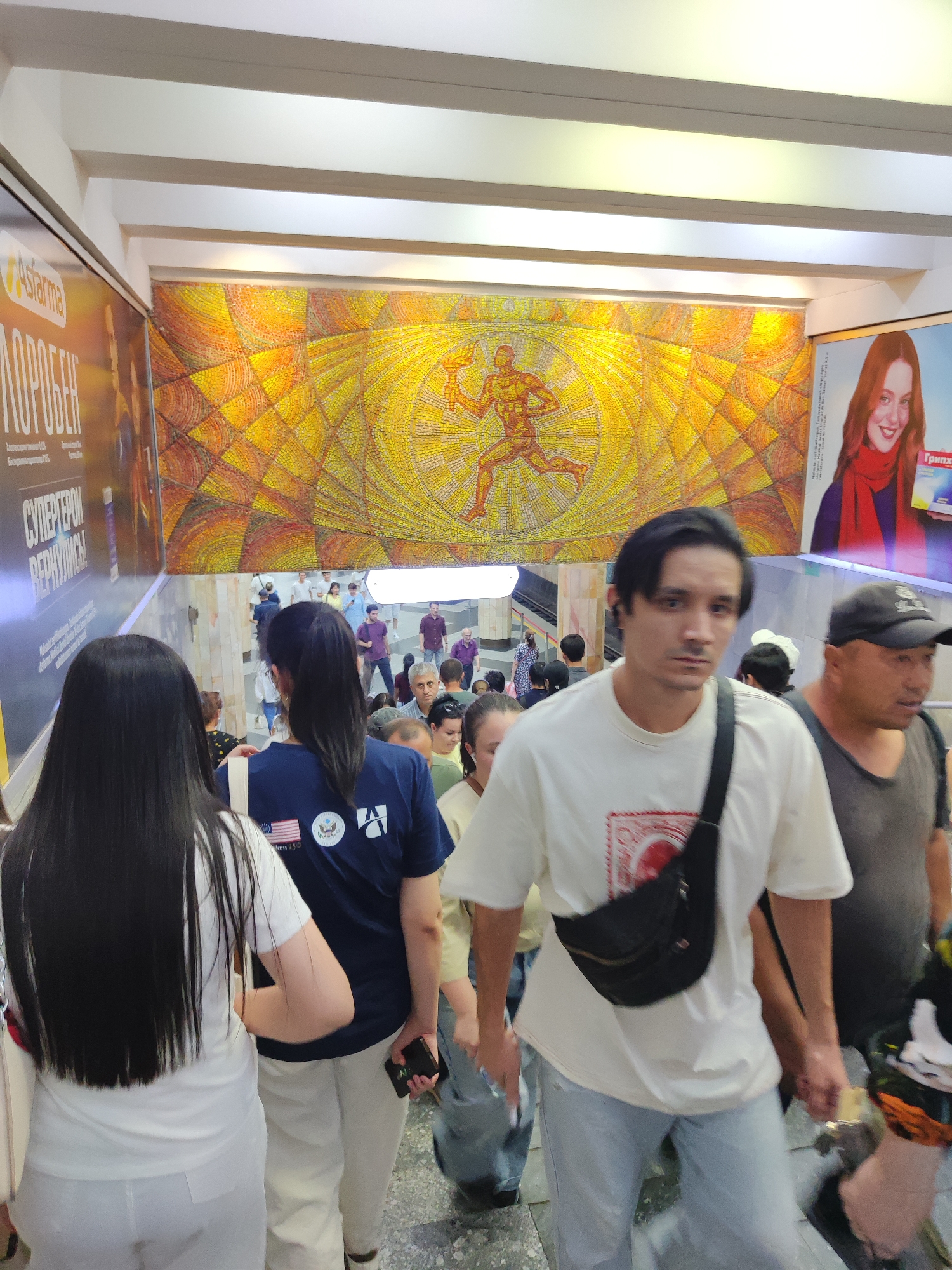The image depicts a scene at a subway station in Tashkent, Uzbekistan, likely on an escalator or staircase leading down from the platform. A group of people are visible, most of them appearing to be commuters. In the foreground, a man with dark hair is looking towards the camera, wearing a white t-shirt and a black fanny pack. To his left, a woman with long black hair is seen from the back, wearing a white top. To the right of the man, an older man wearing a black baseball cap and a grey t-shirt is also present, looking to his right. Further down the stairs, more people are ascending and descending, some with their backs to the camera.
The environment features a distinctive mosaic artwork on the wall behind the people, featuring a stylized figure in motion against a vibrant yellow and orange background. Advertisements are also visible on the left and right walls, including one with a woman's face and text. The lighting suggests an indoor, underground setting. The overall impression is of a bustling public transportation hub.