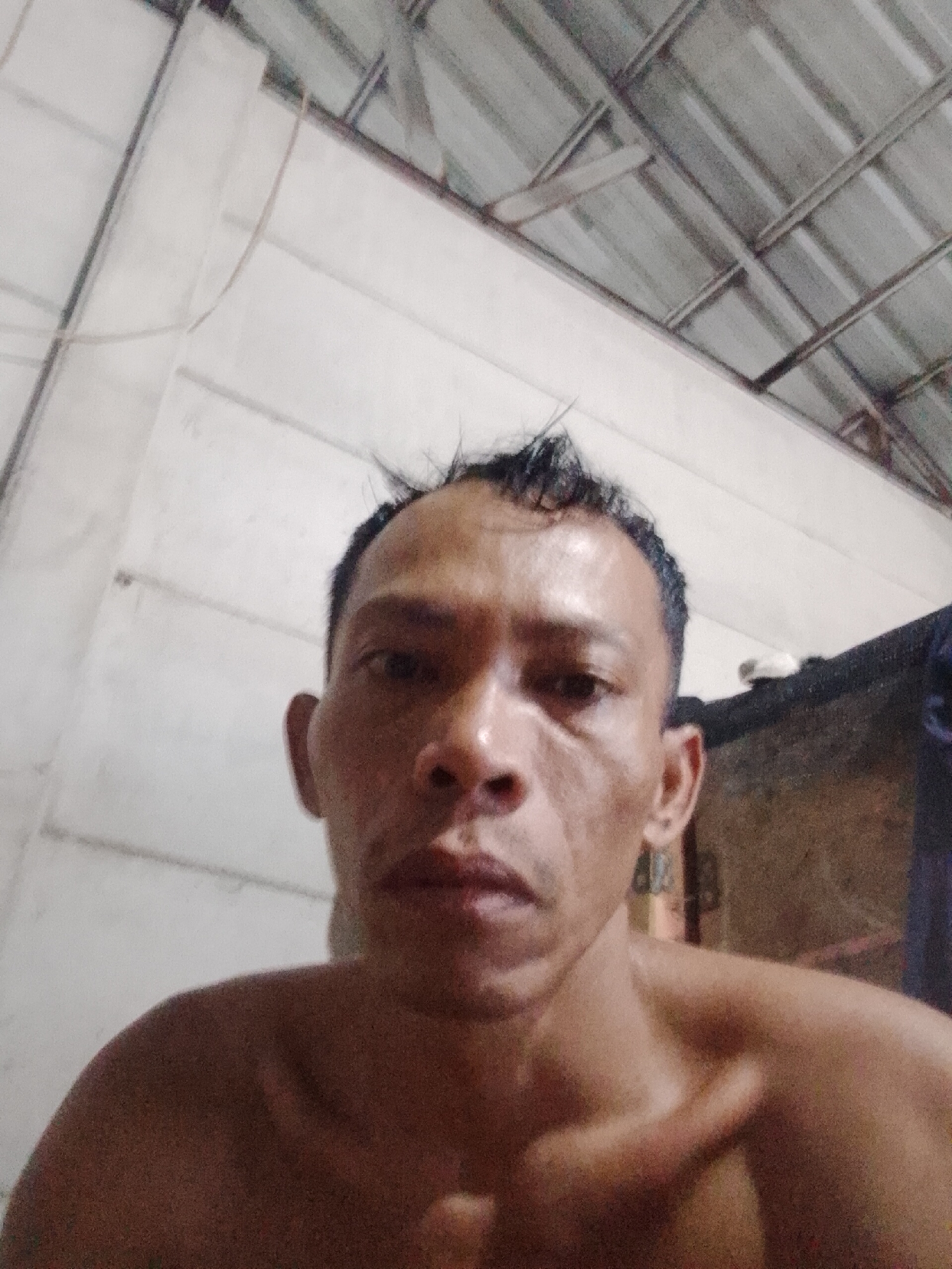 A close-up shot of a shirtless man with short, dark hair is looking at the camera. He appears to be inside, as evidenced by the white wall and the ceiling that resembles a corrugated metal roof with support beams. The exact location within Bukit Ketok, Indonesia cannot be determined from the image alone.