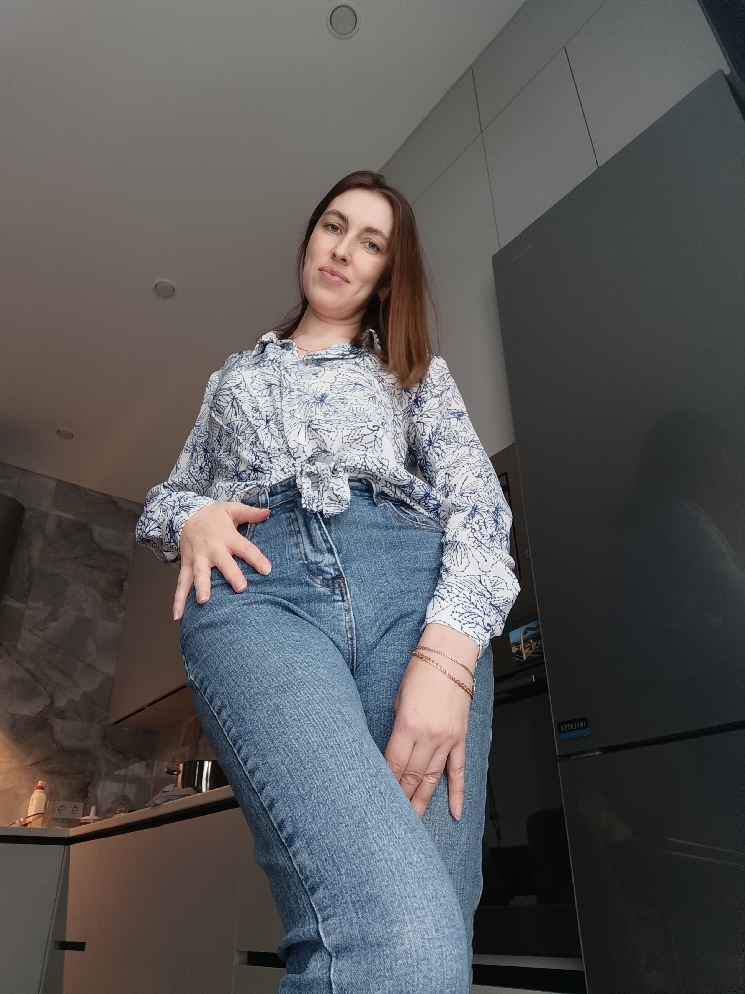 A woman stands in a modern kitchen. She wears a patterned blouse tied at the waist and blue jeans. She has light brown hair. Her right hand rests on her hip, and her left hand is near her leg. The kitchen features gray cabinets, a stainless steel refrigerator, and a light-colored countertop with kitchen utensils visible. The location is Chaikovsky, Russia.