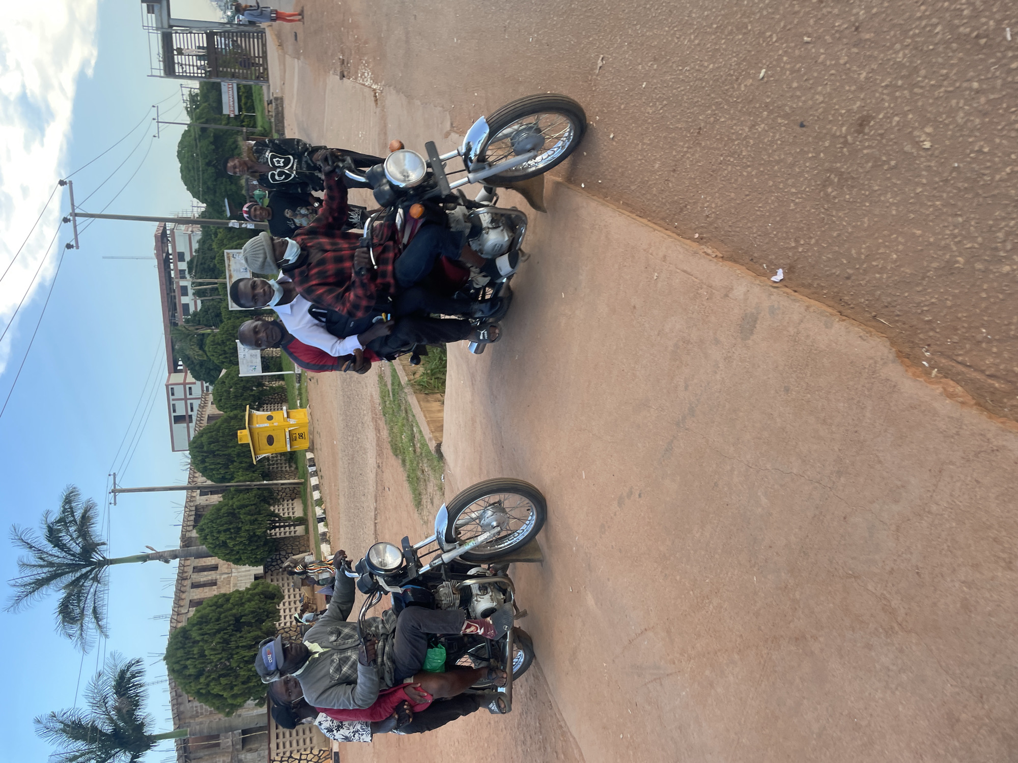 In Jos, Nigeria, two motorcycles are carrying multiple passengers each, traveling along a paved road with a concrete median. The motorcycle on the left carries three individuals, with the driver wearing a grey jacket and a dark cap, and two passengers behind him. The motorcycle on the right is also carrying three individuals, with the driver in a red and black plaid shirt and a grey beanie, and two passengers behind him. In the background, there are buildings under construction, trees, and utility poles. The sky is partly cloudy.