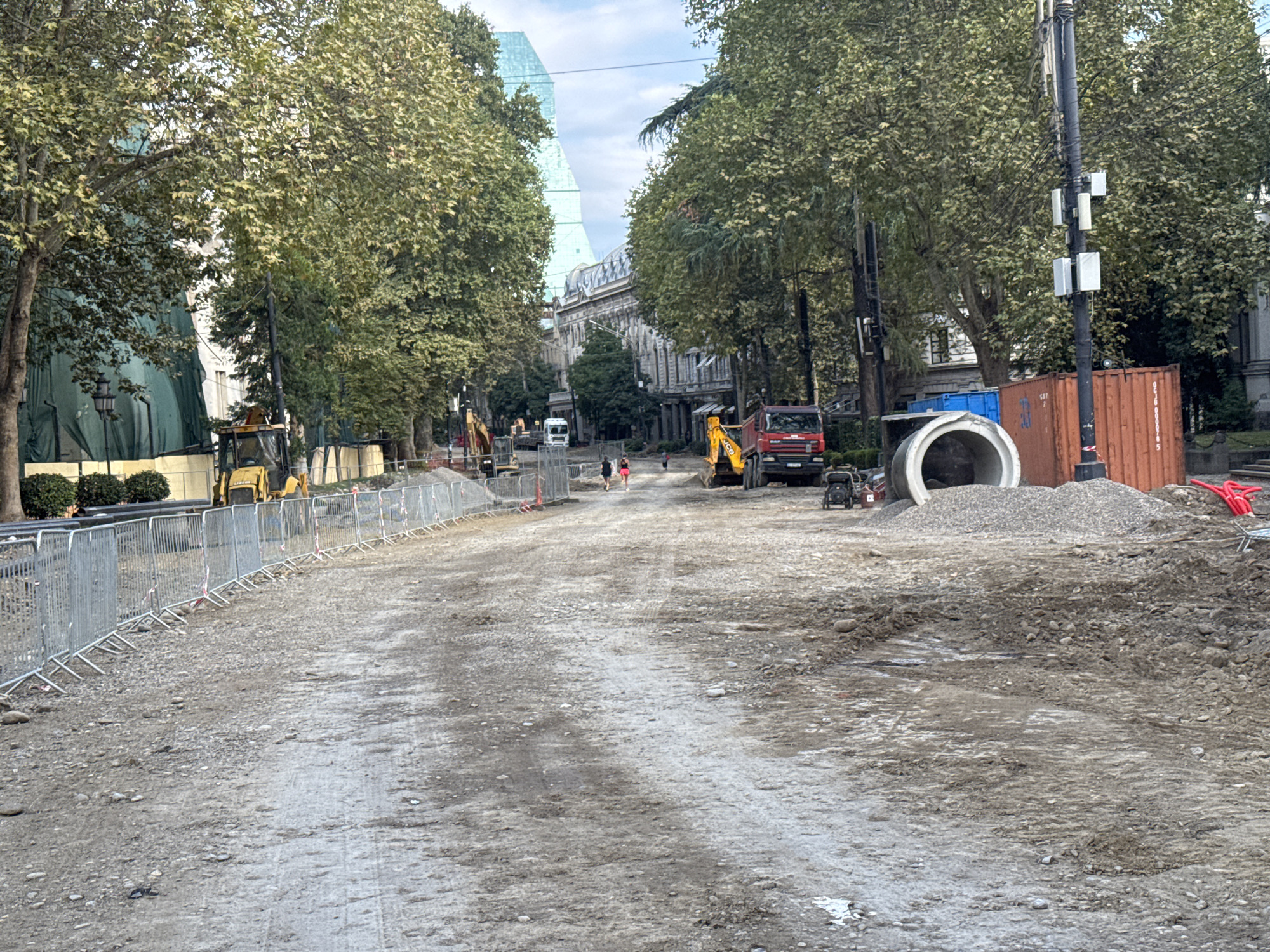 The image depicts a construction site on Rustaveli Avenue in Tbilisi, Georgia. In the foreground, a dirt and gravel road is visible, with temporary metal barriers lining the left side. Construction vehicles, including a yellow excavator and a red dump truck, are present. Large concrete pipes and a red shipping container are situated on the right side of the road. Several people, appearing to be workers or passersby, are walking in the middle distance. The background shows mature trees lining the avenue, with historic buildings and a modern glass skyscraper visible behind them. The scene suggests ongoing infrastructure work in a central urban area.